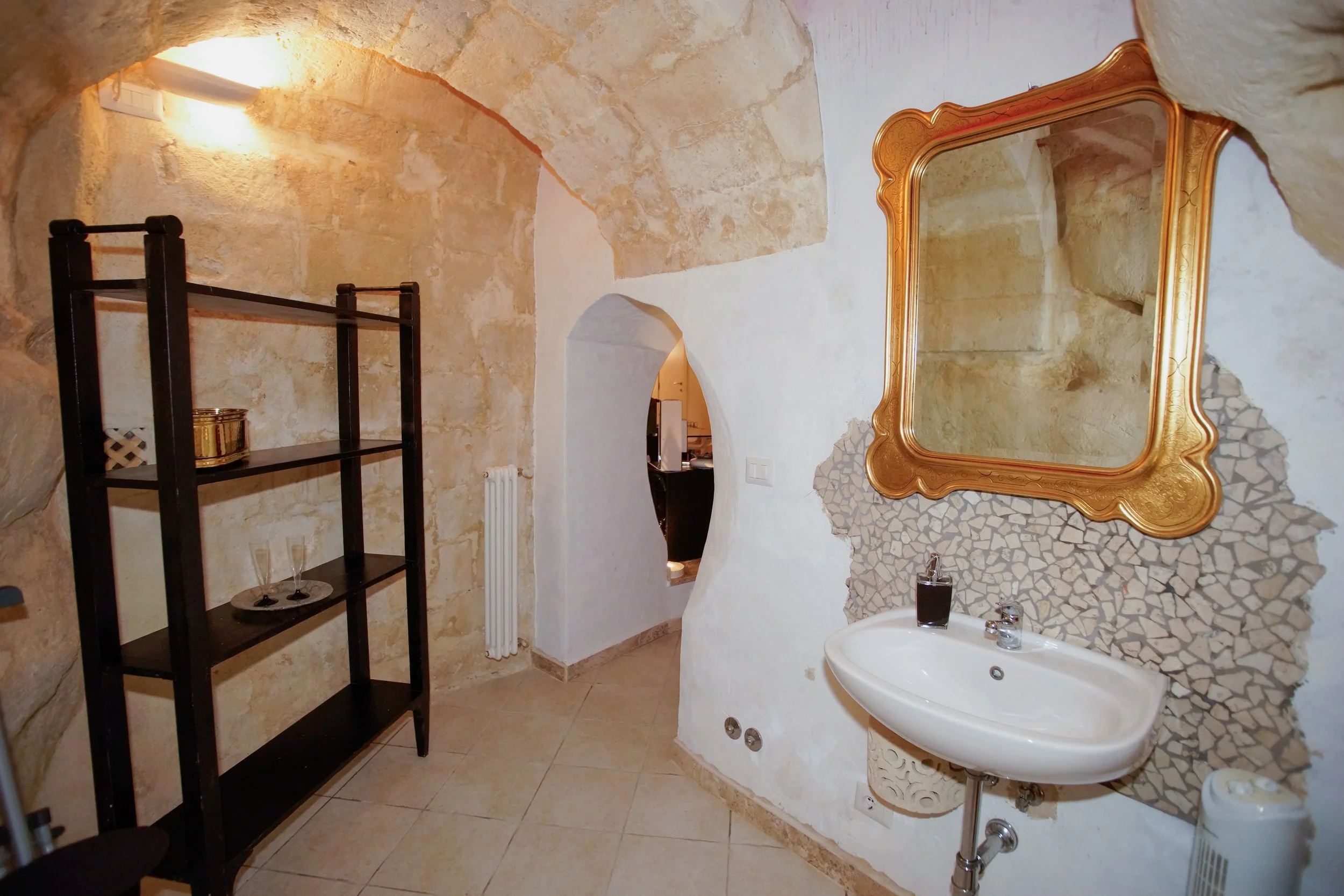 Bagno con pareti in pietra e mosaico, grande specchio dorato, lavabo con sapone, scaffale nero con oggetti decorativi e bicchieri, archi in pietra, pavimento in piastrelle beige.