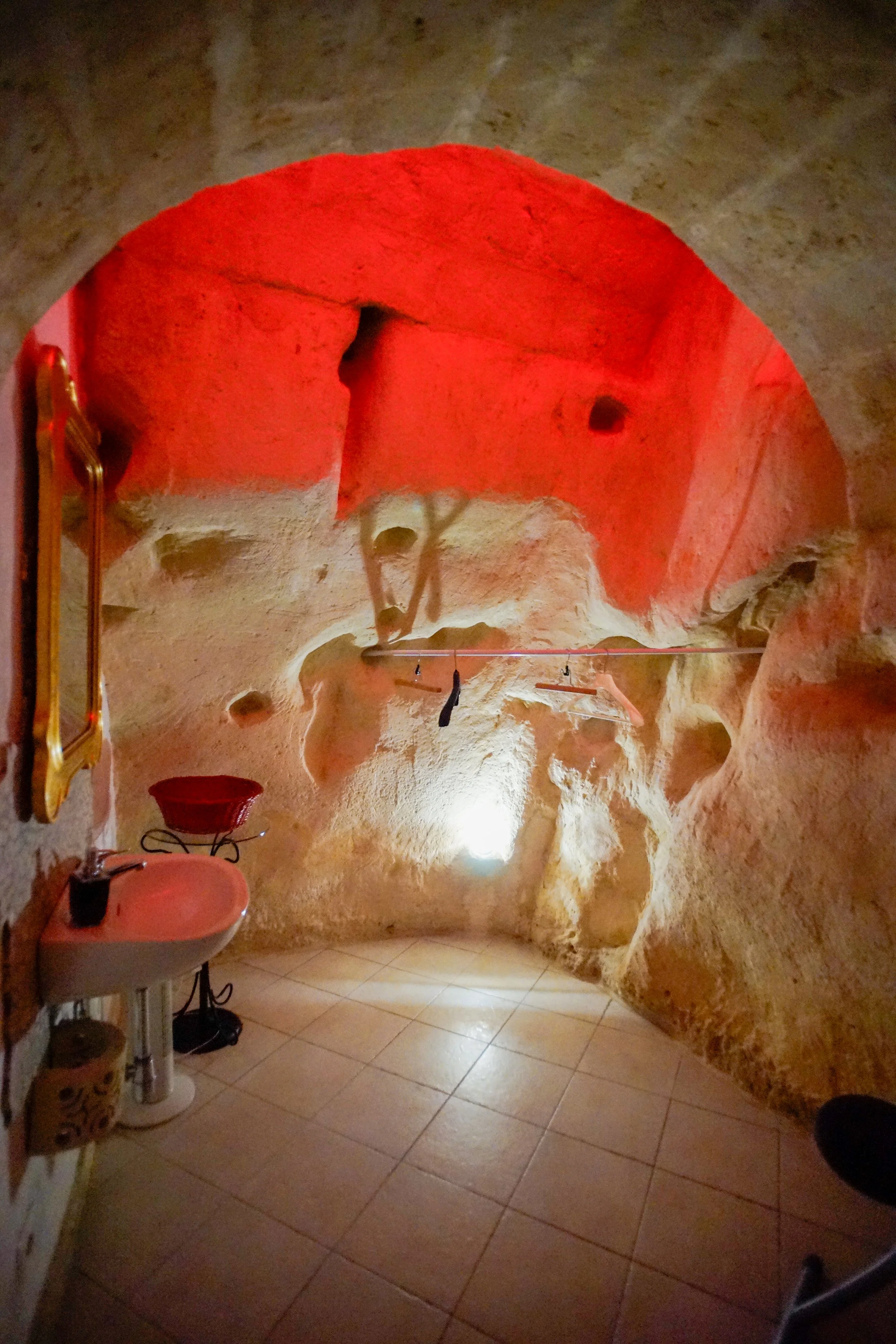 Interno di una grotta con pareti di rocce sabbiose, illuminata da luci calde, con lavabo, specchio e oggetti appesi alla parete.