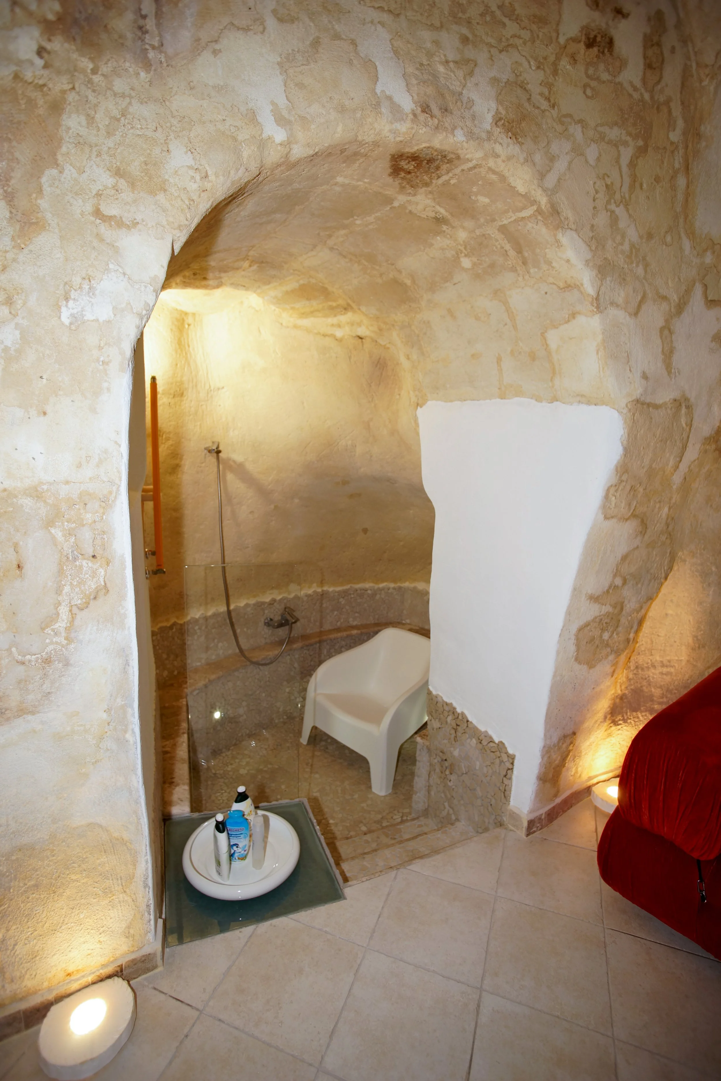 Bagno in una grotta con pareti in pietra, con doccia, sedia bianca e prodotti per l'igiene, illuminato da luci calde.