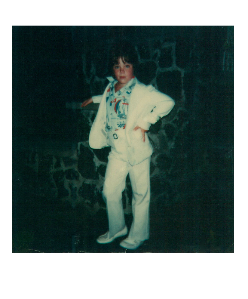 Child dressed in a white suit with a colorful shirt, standing against a dark stone wall with a confident pose.