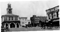 A historical black and white photo of a town square with a clock tower, surrounding buildings, and horse-drawn carriages. Fleishman's can be seen on the right hand side of the image. Lineage of Thompson's, a Men’s clothing store in Wilmington