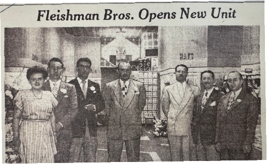 Group of seven people standing in front of a store, celebrating the opening of a new unit, with a banner reading 'Fleishman Bros. Opens New Unit'. Lineage for Thompson's of Wilmington, a Men’s clothing store in Wilmington NC.