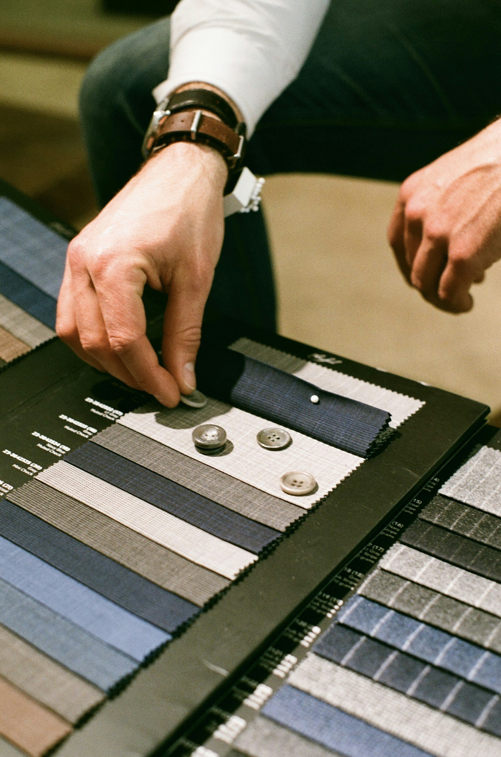 Close-up of a person selecting a fabric swatch from a color palette, with several buttons on the swatch. Tailored clothing for Thompson's of Wilmington, a Men’s clothing store in Wilmington NC.