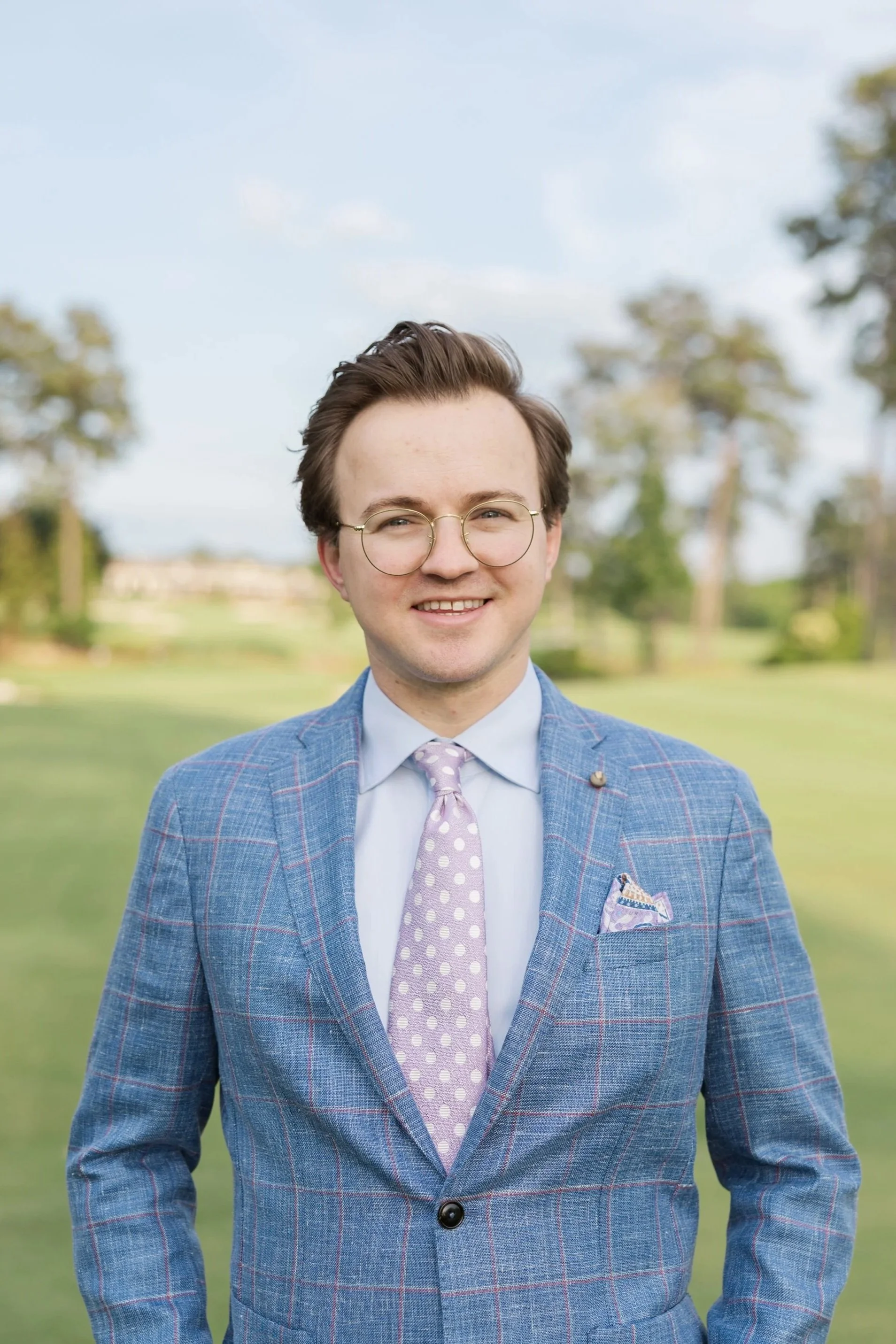 Bryce Thompson with glasses smiling outdoors in a park with green grass and trees, wearing a blue checkered custom sport coat, tailored white shirt, and light purple polka dot tie. Owner of Thompson's, a Men’s clothing store in Wilmington