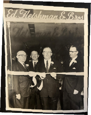 Four men in suits and ties at a ribbon-cutting ceremony, with a banner reading 'Ed. Fleishman & Bros.' in the background. Lineage of Thompson's of Wilmington, a Men’s clothing store in Wilmington NC.