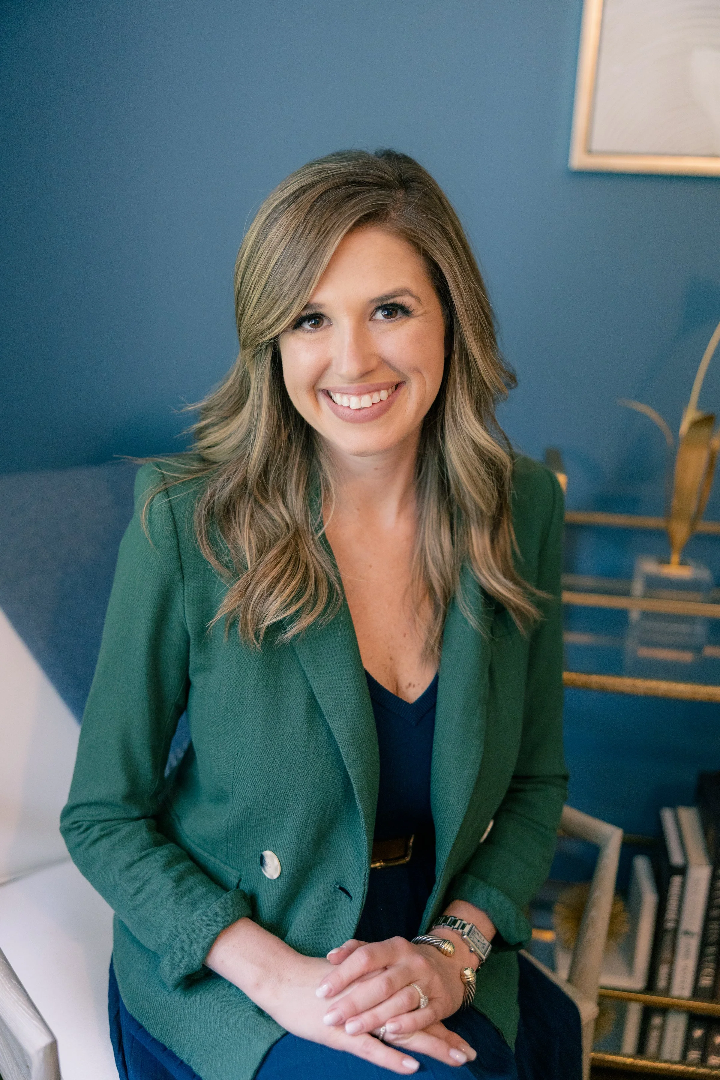 Maggie Thompson with shoulder-length wavy blonde hair wearing a green blazer and navy top sitting and smiling in a room with a blue wall background and a gold bookshelf. Owner of Thompson's of Wilmington, a Men’s clothing store in Wilmington NC.