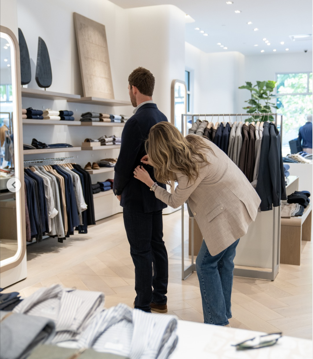 Maggie Thompson fitting a man's sport coat in a clothing store, with racks of clothes and shelves of fabric behind them. Owner for Thompson's of Wilmington, a Men’s clothing store in Wilmington NC.