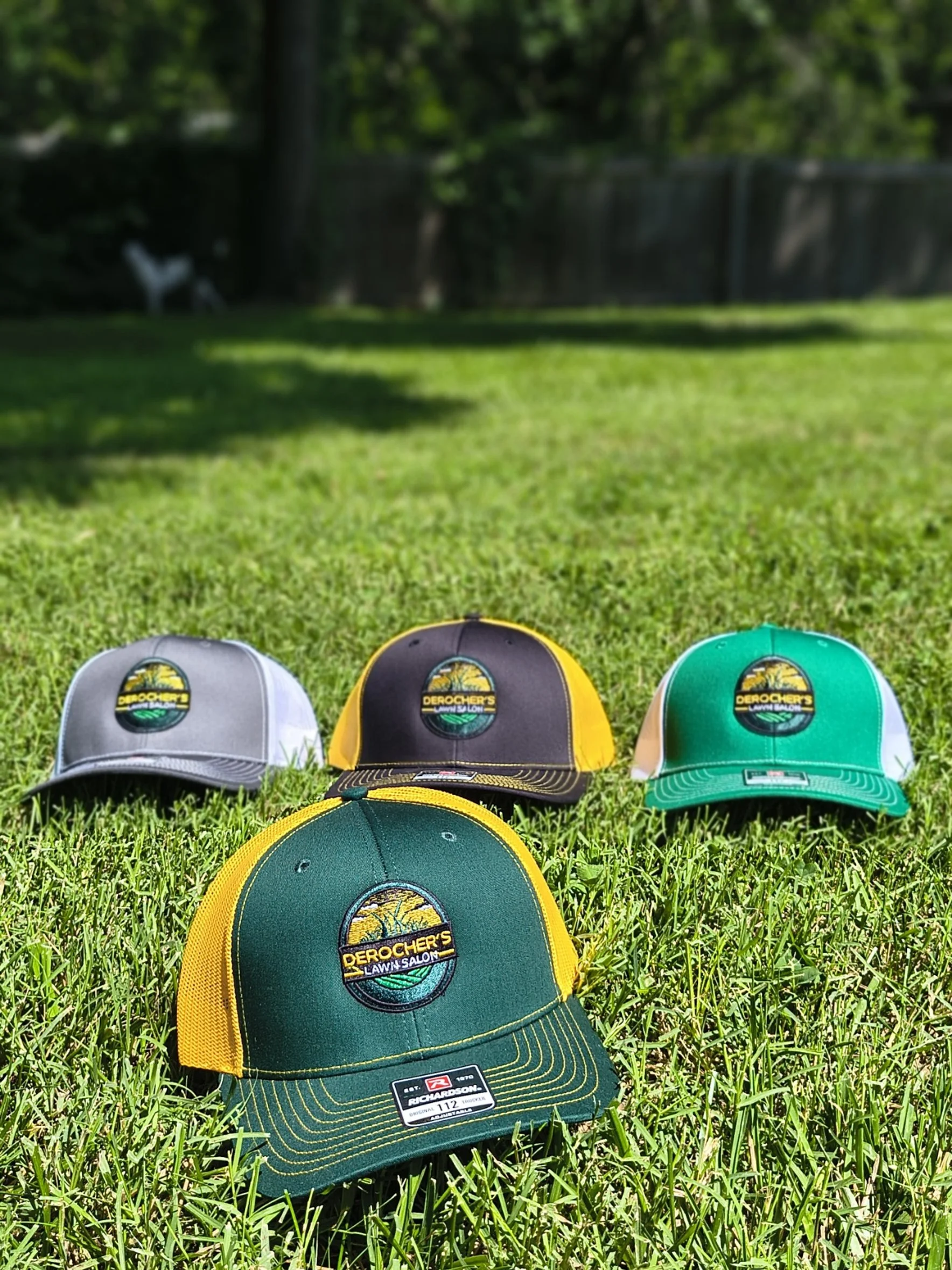 Four baseball caps with logos for Derogher's Lawn Salon on a grassy field, with a blurred background of a wooden fence, trees, and a dog.