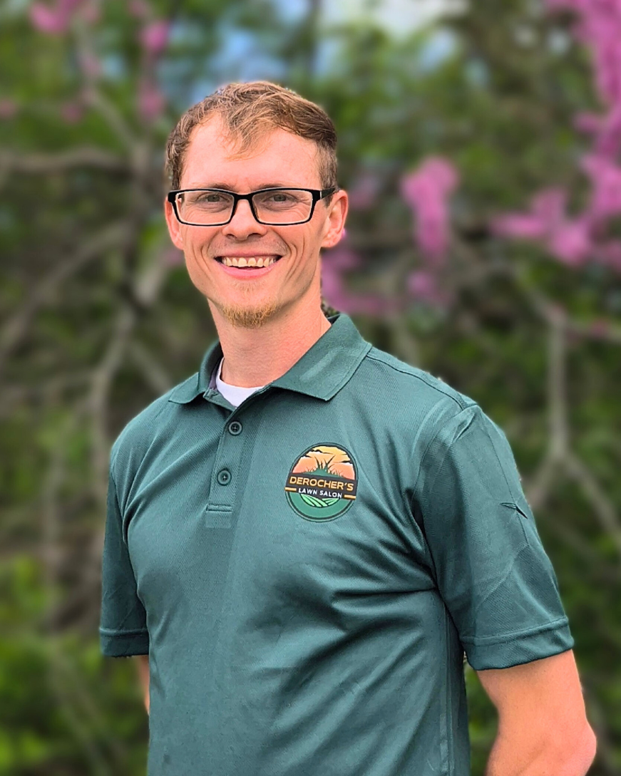 A young man with glasses smiling outdoors, wearing a teal polo shirt with a logo that reads Derocher's Lawn Salon, standing in front of a blurred background of green foliage and pink flowers.
