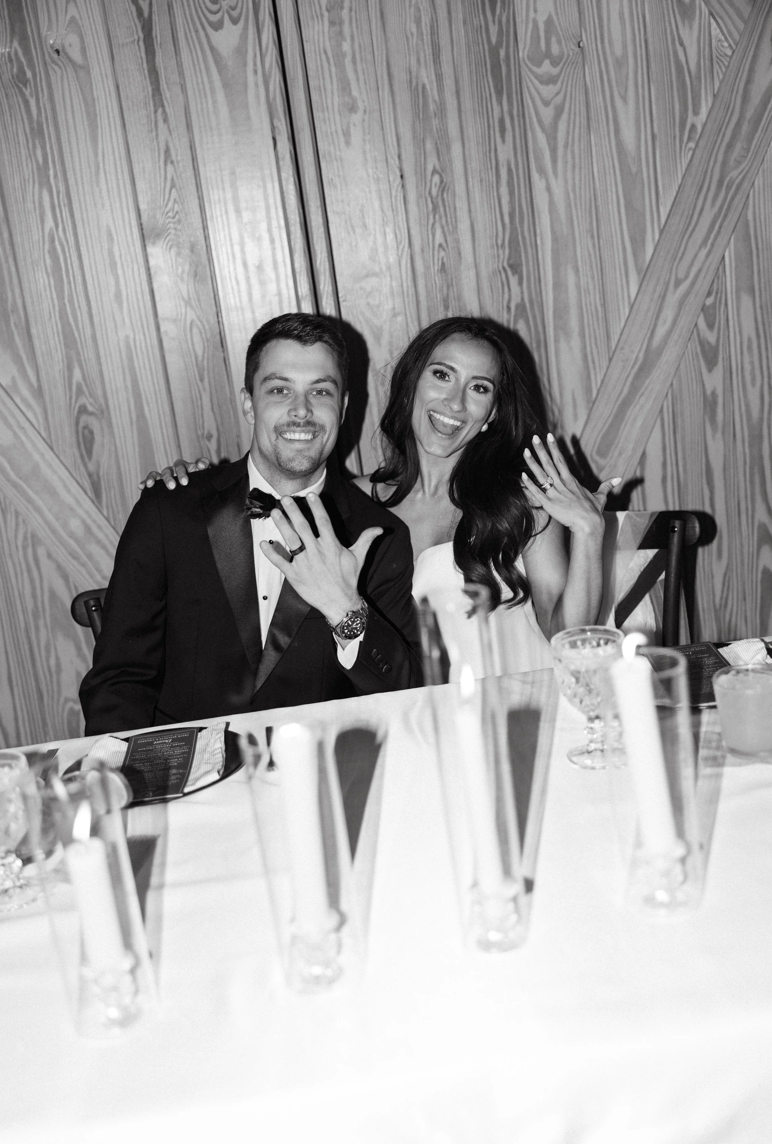Black and white photo of a happy couple at a wedding reception, sitting at a decorated table with drinks, showing their wedding rings.