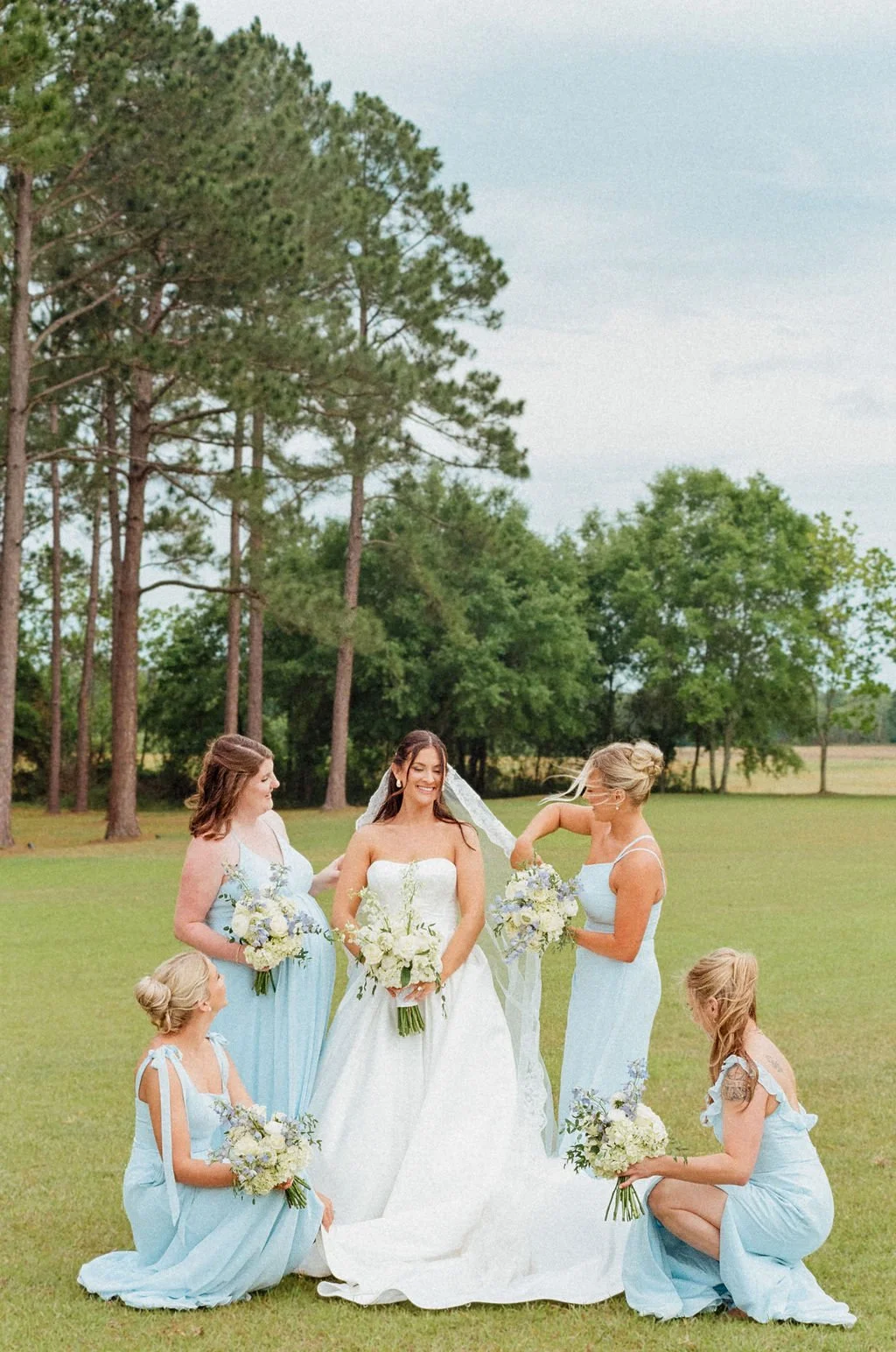A bride with four bridesmaids and two young flower girls in a grassy field with trees, celebrating an outdoor wedding.