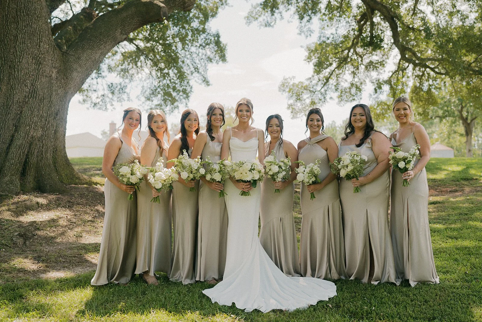 A group of nine women in matching beige dresses standing outdoors in front of large trees, holding bouquets of white flowers, with a grass field and houses in the background, on a sunny day.
