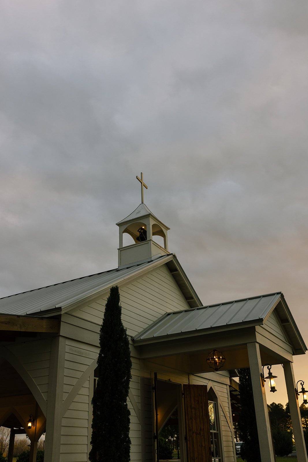 A white church with a steeple and a cross on top, under a cloudy sky, with hanging lanterns near the entrance.