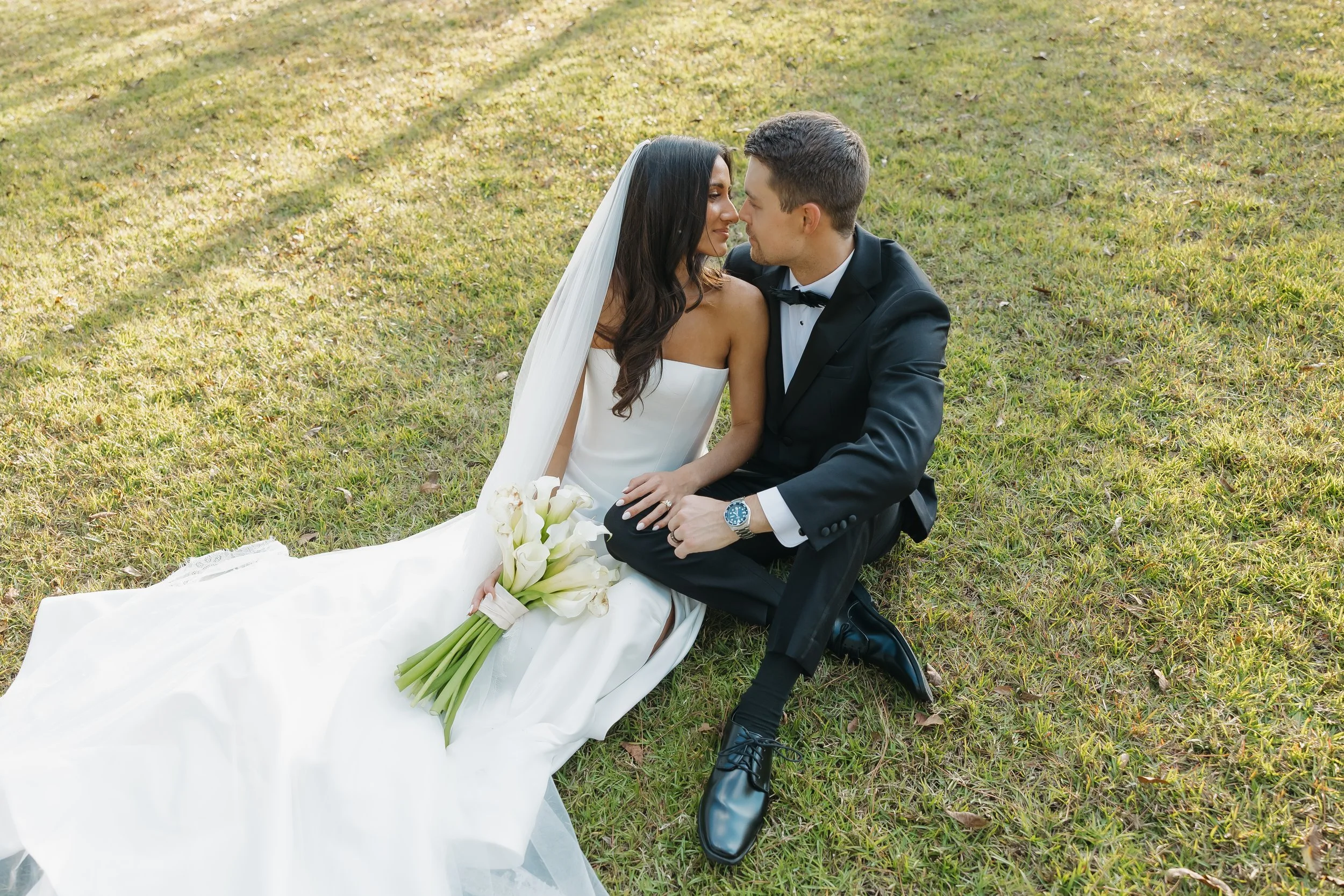 A bride and groom sit closely on the grass, facing each other, during their wedding. The bride wears a white strapless wedding gown with a veil and holds a bouquet of calla lilies. The groom wears a black tuxedo with a bow tie and a watch.