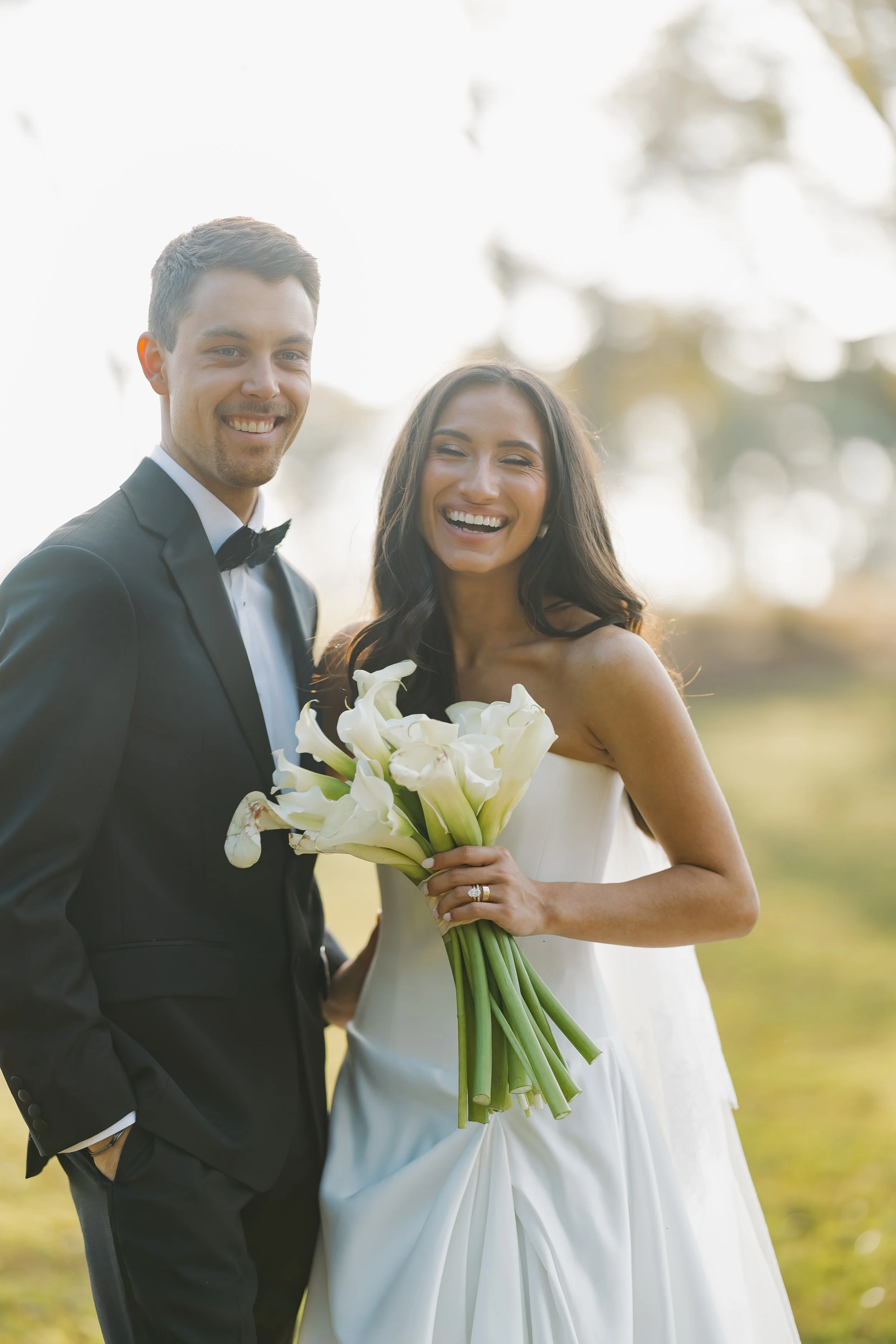 A newlywed couple smiling outdoors, with the groom in a black tuxedo and the bride in a strapless white wedding dress holding a bouquet of white calla lilies.