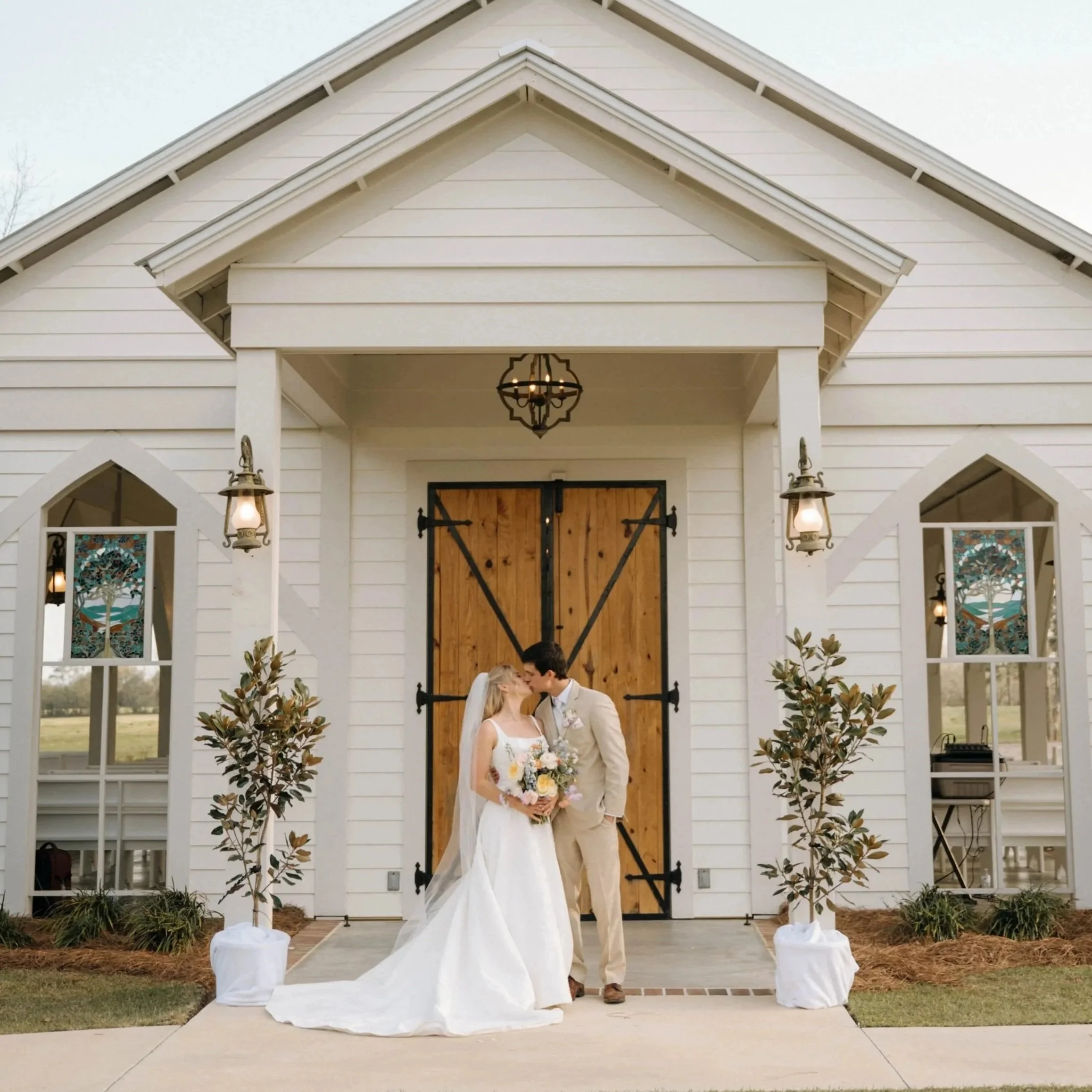 A bride and groom are sharing a kiss outside a white wedding chapel with wooden doors, flanked by two potted trees and decorative lanterns.