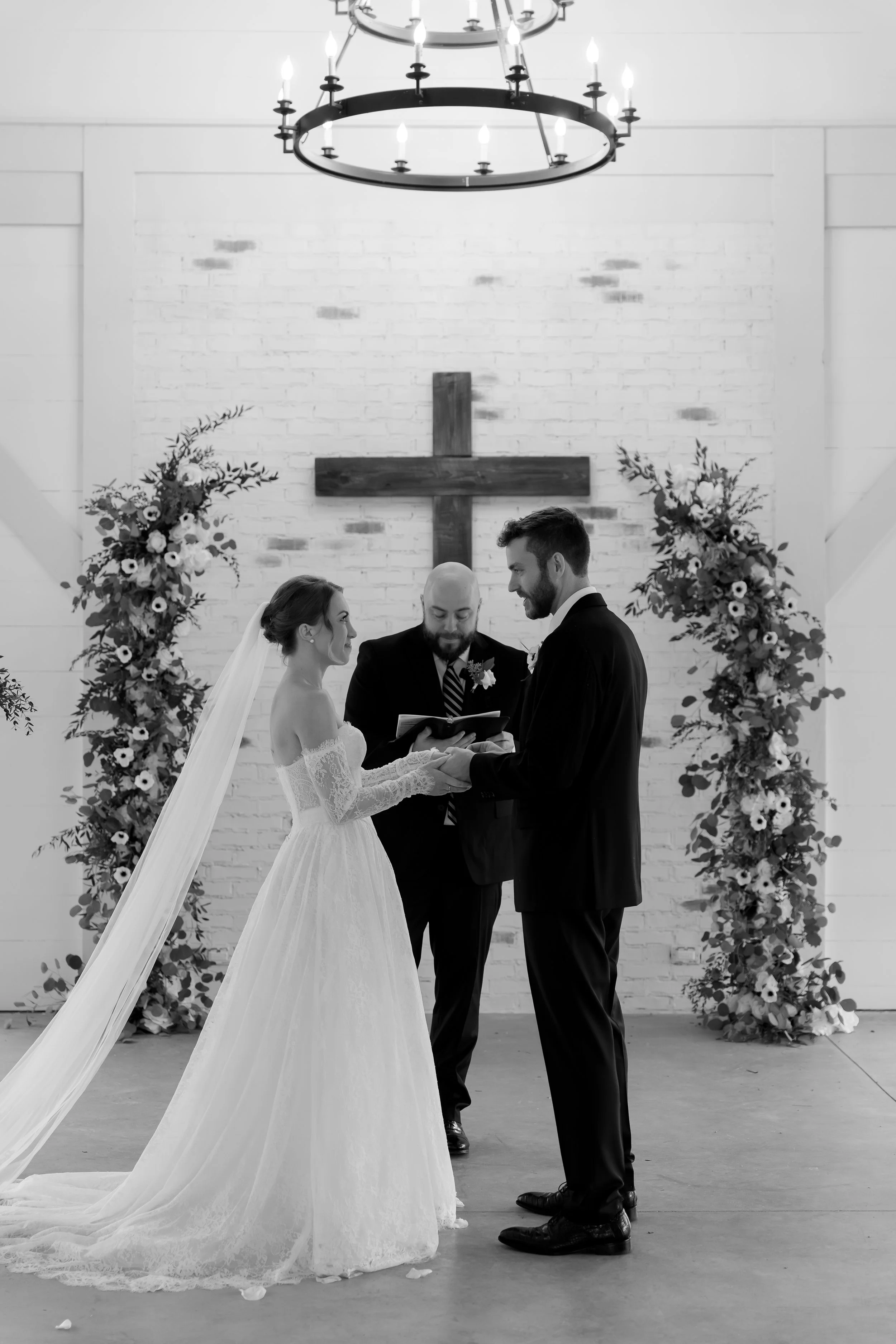 Black and white photo of a wedding ceremony in a church with a bride, groom, and officiant. The bride is in a white wedding dress with a veil, and the groom in a suit. They are holding hands, facing each other, with a cross on the brick wall behind t