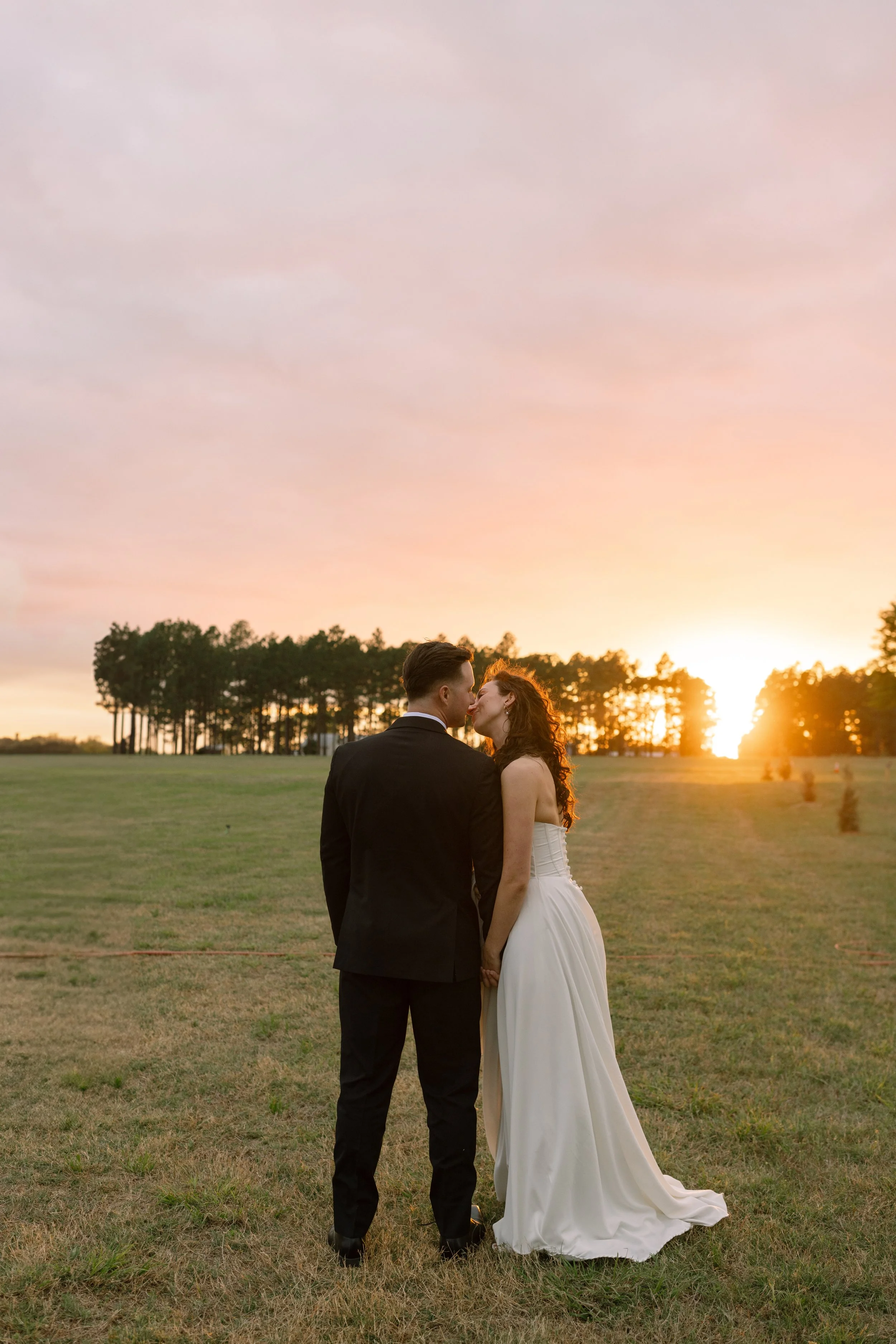 A bride and groom stand close together in a grassy field during sunset, holding hands and about to kiss, with a line of trees in the background.