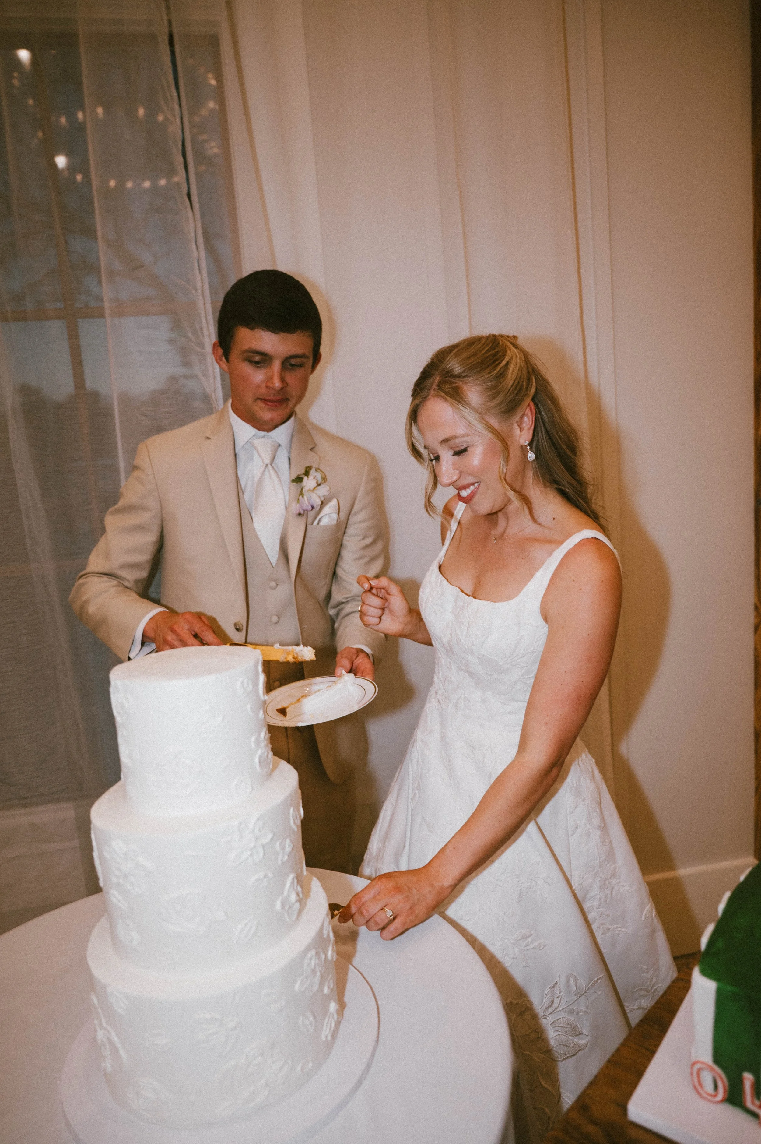 A bride and groom cutting a wedding cake together at their wedding reception. The wedding cake is white with textured, floral-like patterns and three tiers.