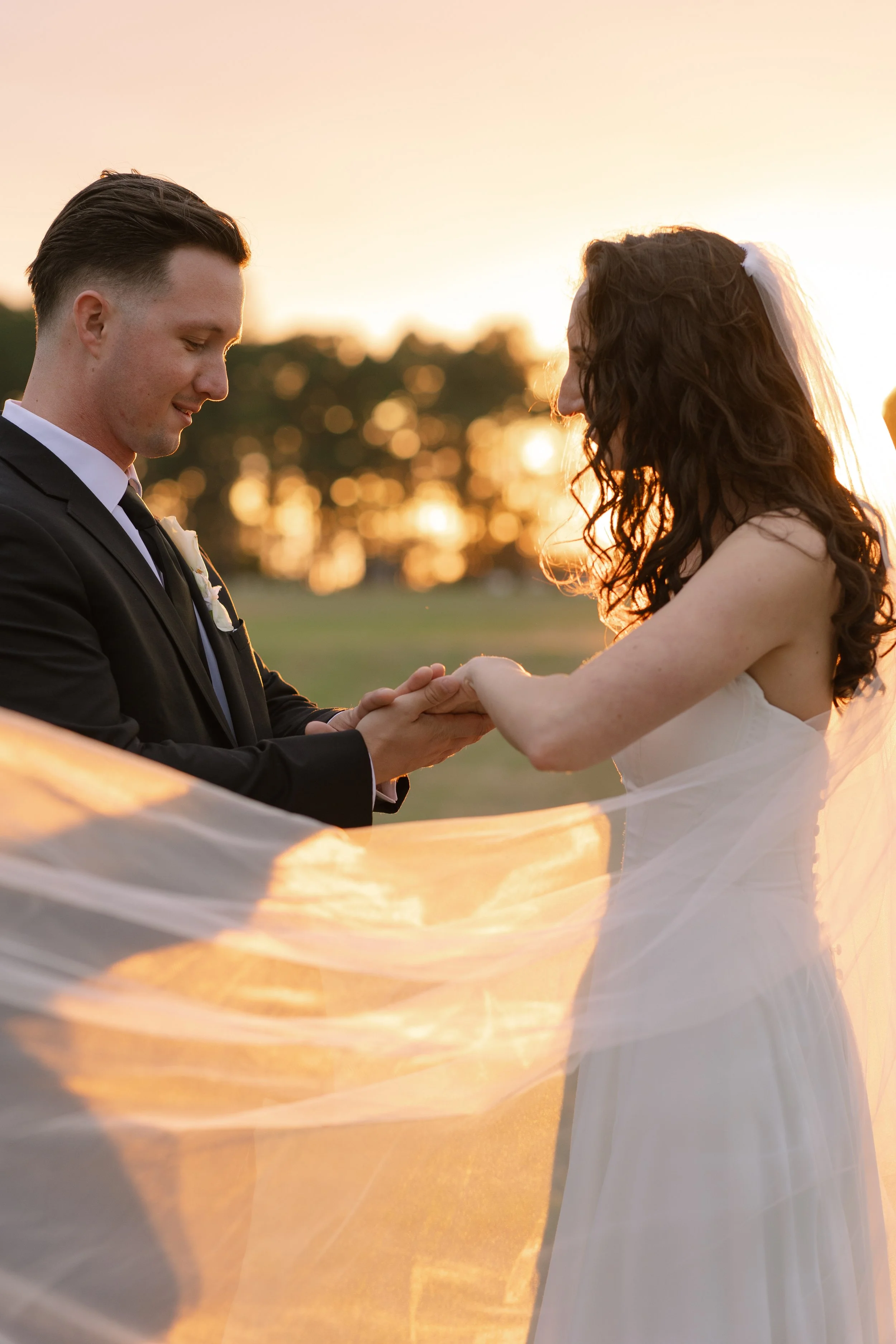 A groom and bride stand close together during a wedding ceremony at sunset, holding hands and smiling at each other, with flowing veil and blurred trees in the background.