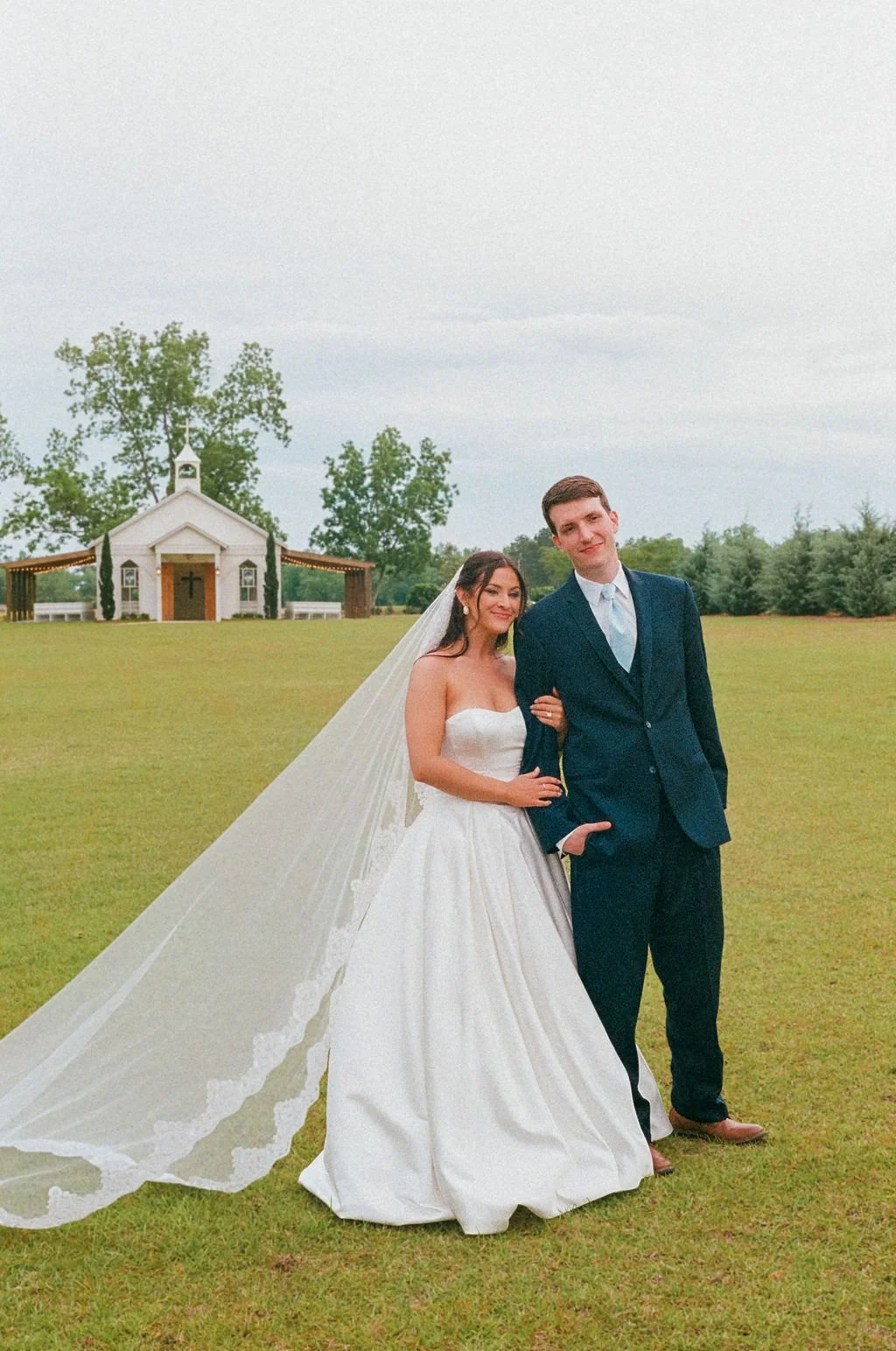 A bride and groom standing on a grassy field with a small chapel in the background. The bride is wearing a strapless white wedding dress and a long veil, and the groom is dressed in a dark blue suit. They are smiling and holding each other.