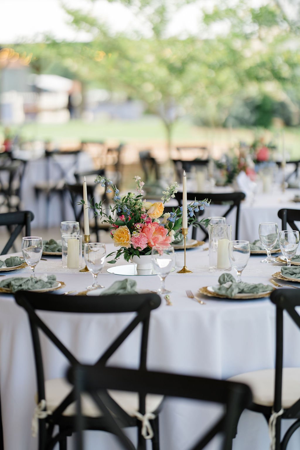 Elegant outdoor wedding reception table with floral centerpiece, candles, glassware, and gold-rimmed plates, set against lush green trees.