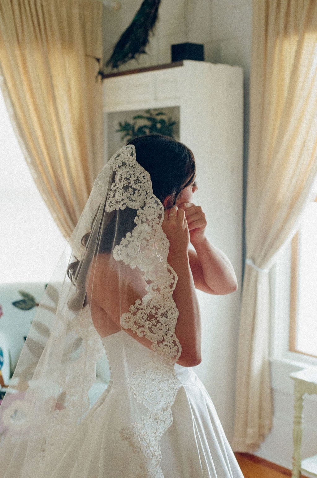 A bride with a lace veil on her head, adjusting her earrings in a room with cream-colored curtains.