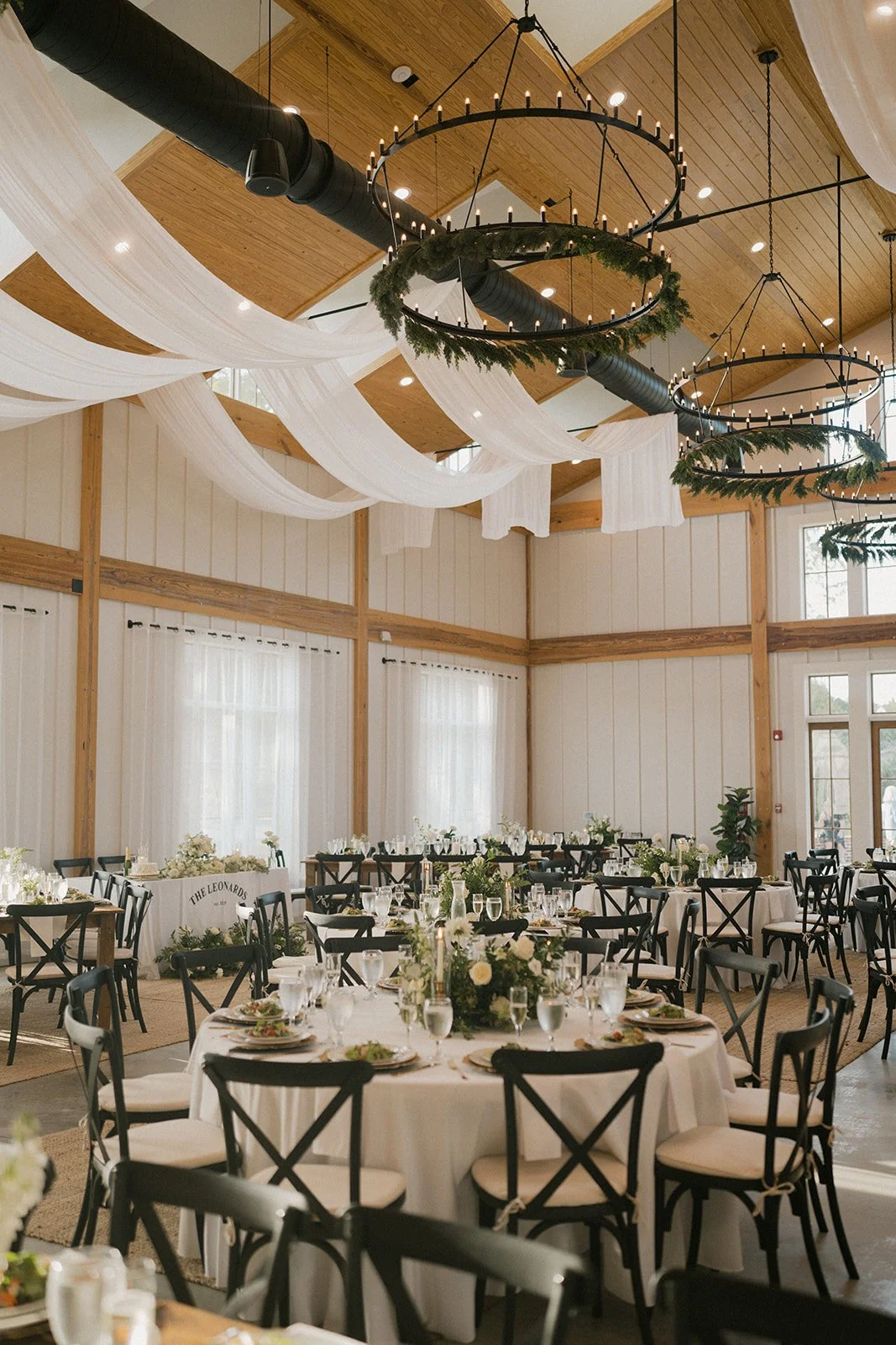 Event hall decorated with white drapes, large circular chandeliers with candles, wooden ceiling, and round tables with white tablecloths, floral centerpieces, and place settings, suitable for a wedding or celebration.