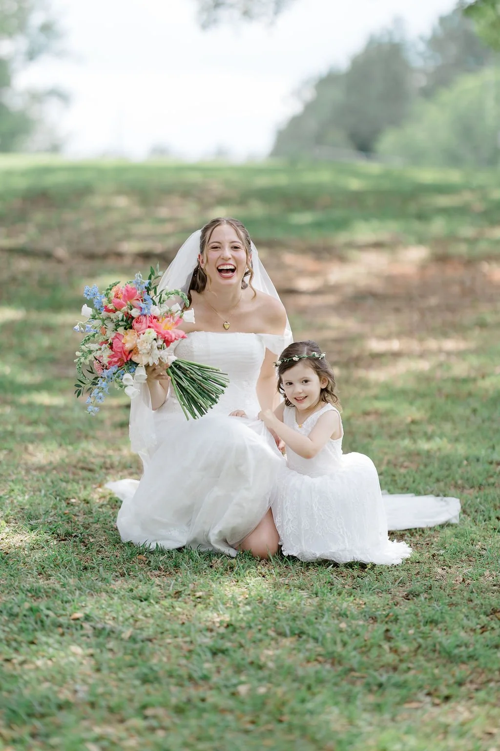 A bride in a white wedding dress sitting on grass, holding a colorful bouquet of flowers, and smiling, next to a young girl in a white dress with a floral headband, outdoors in a park with green trees and grass in the background.