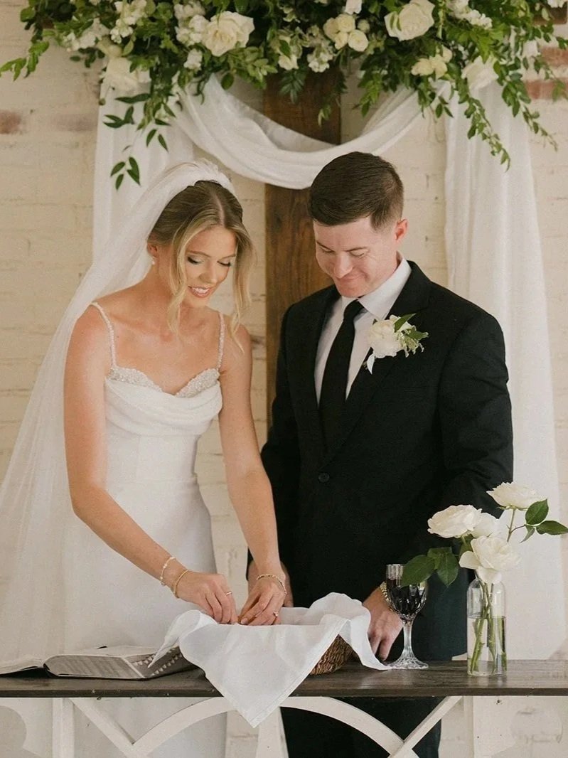 Bride and groom taking communion at their wedding ceremony in a decorated venue with white flowers and fabric backdrop.