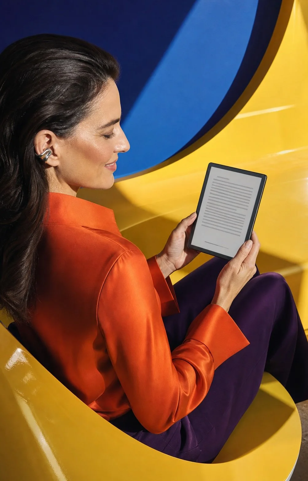 Woman in an orange silk blouse reading an e-reader against a glossy yellow and cobalt-blue setting.