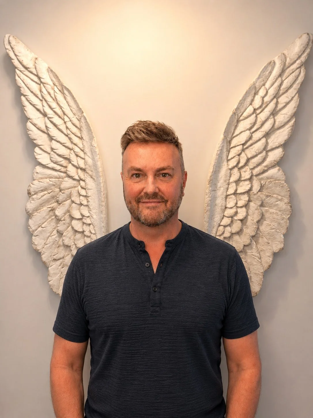 Lewis Mabee standing in front of angel wings, smiling warmly and making direct eye contact, representing over 34 years of conversations, guidance, and connection with clients across generations.