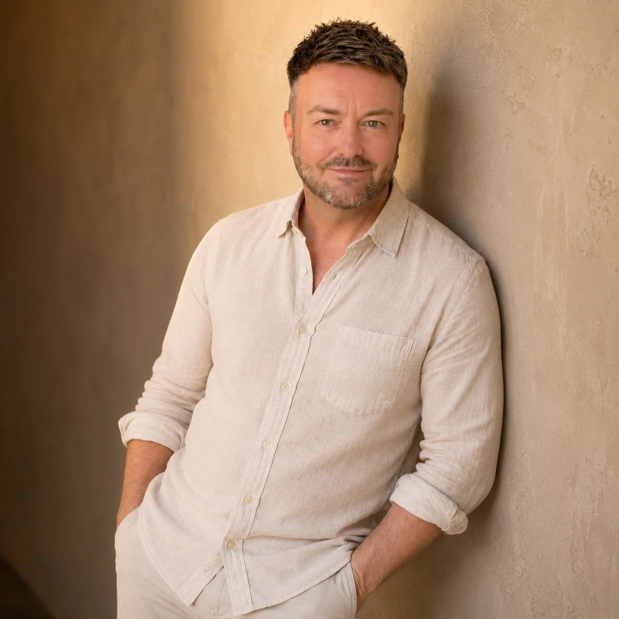 Lewis Mabee leaning casually against a warm neutral wall with a welcoming smile and direct eye contact, creating a relaxed professional portrait that invites visitors to get in touch.