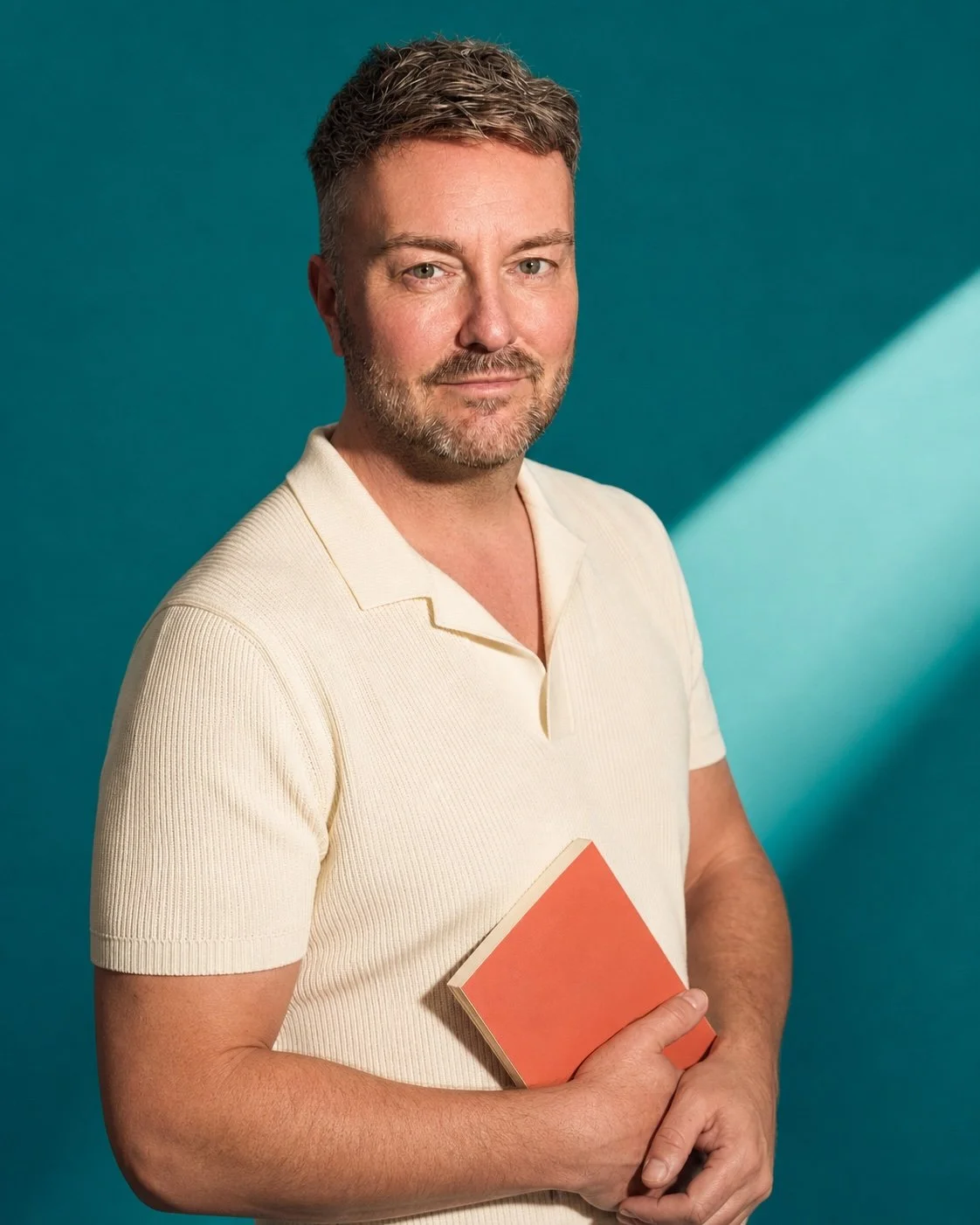 Lewis Mabee holding a coral paperback while wearing an ivory knit shirt against a bright teal studio background.