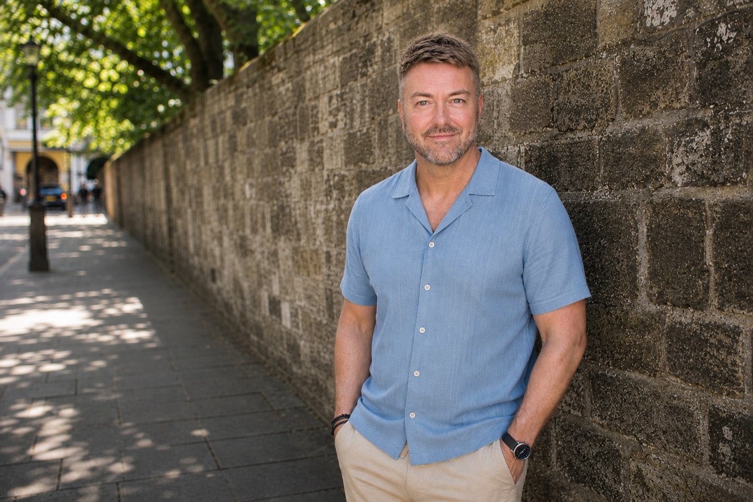 Lewis Mabee is wearing a light blue short-sleeved shirt and beige pants, standing outdoors against a stone wall, with trees and a city street in the background.
