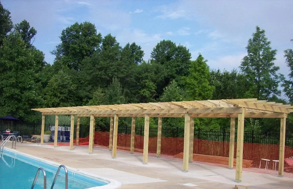 Outdoor swimming pool area with a wooden pergola under construction, surrounded by green trees and a partly cloudy sky.