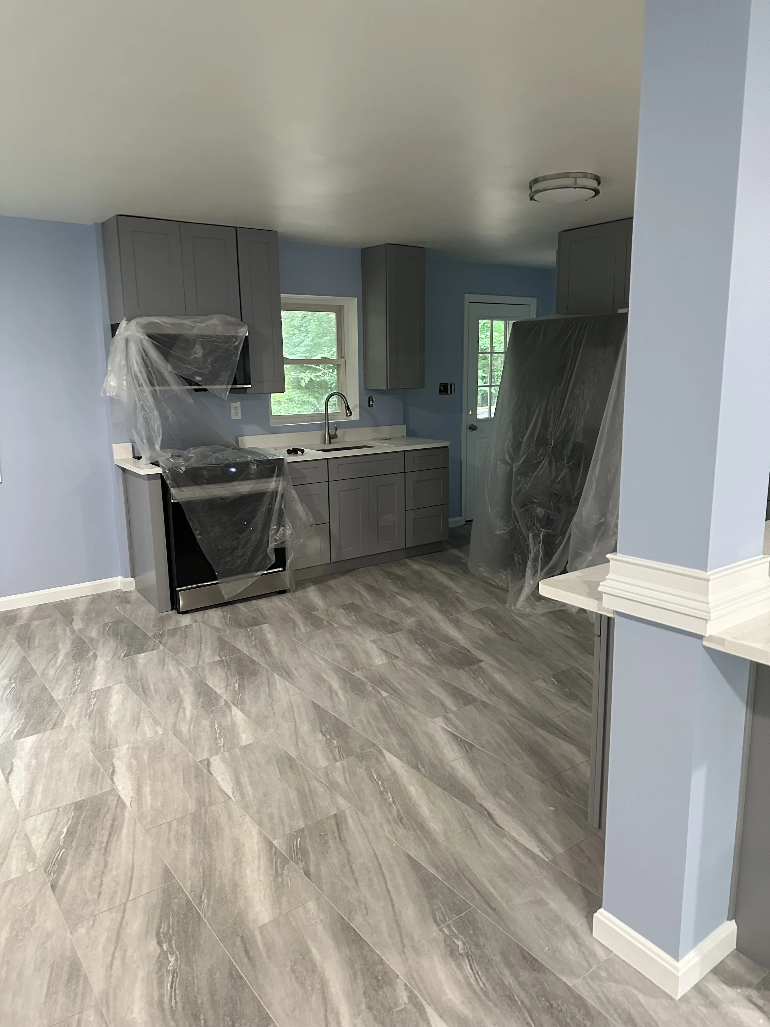 Newly renovated kitchen with blue walls, gray cabinets, and wood-look tile flooring. Plastic protective coverings on appliances and new fixtures, with a window over the sink and a door leading outside.
