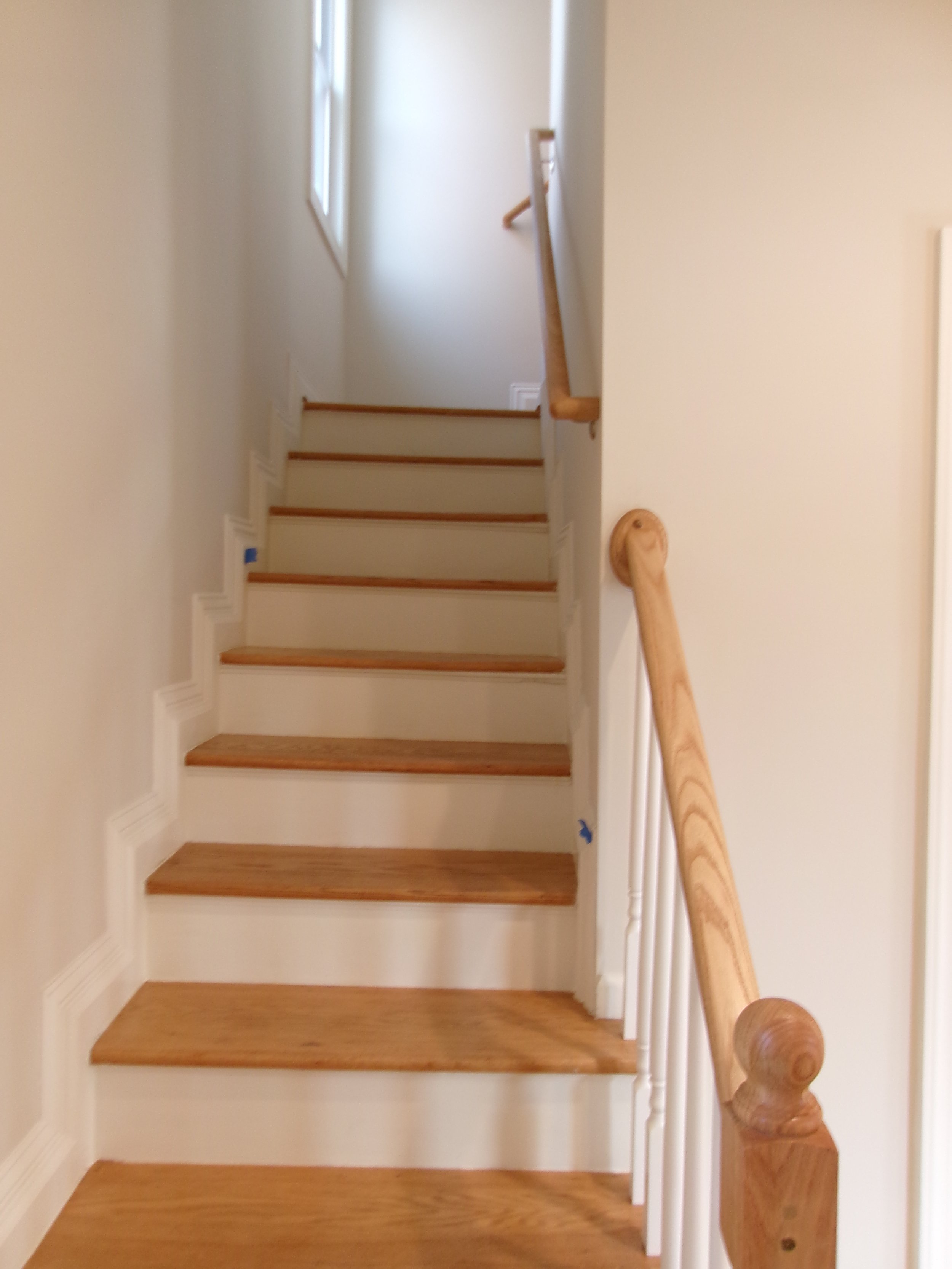 Interior view of a staircase with wooden steps, white risers, wooden handrail on the right, and a window at the top providing natural light.