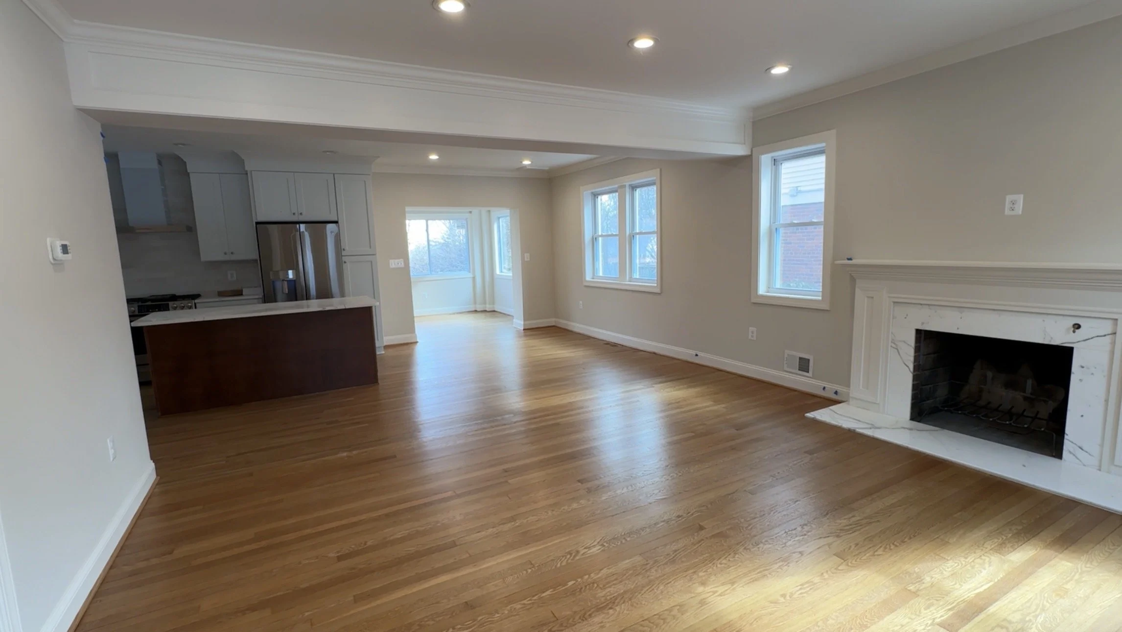 Empty living room with hardwood floors, a fireplace with white mantel, and multiple windows providing natural light. An open kitchen with a small island is visible in the background.