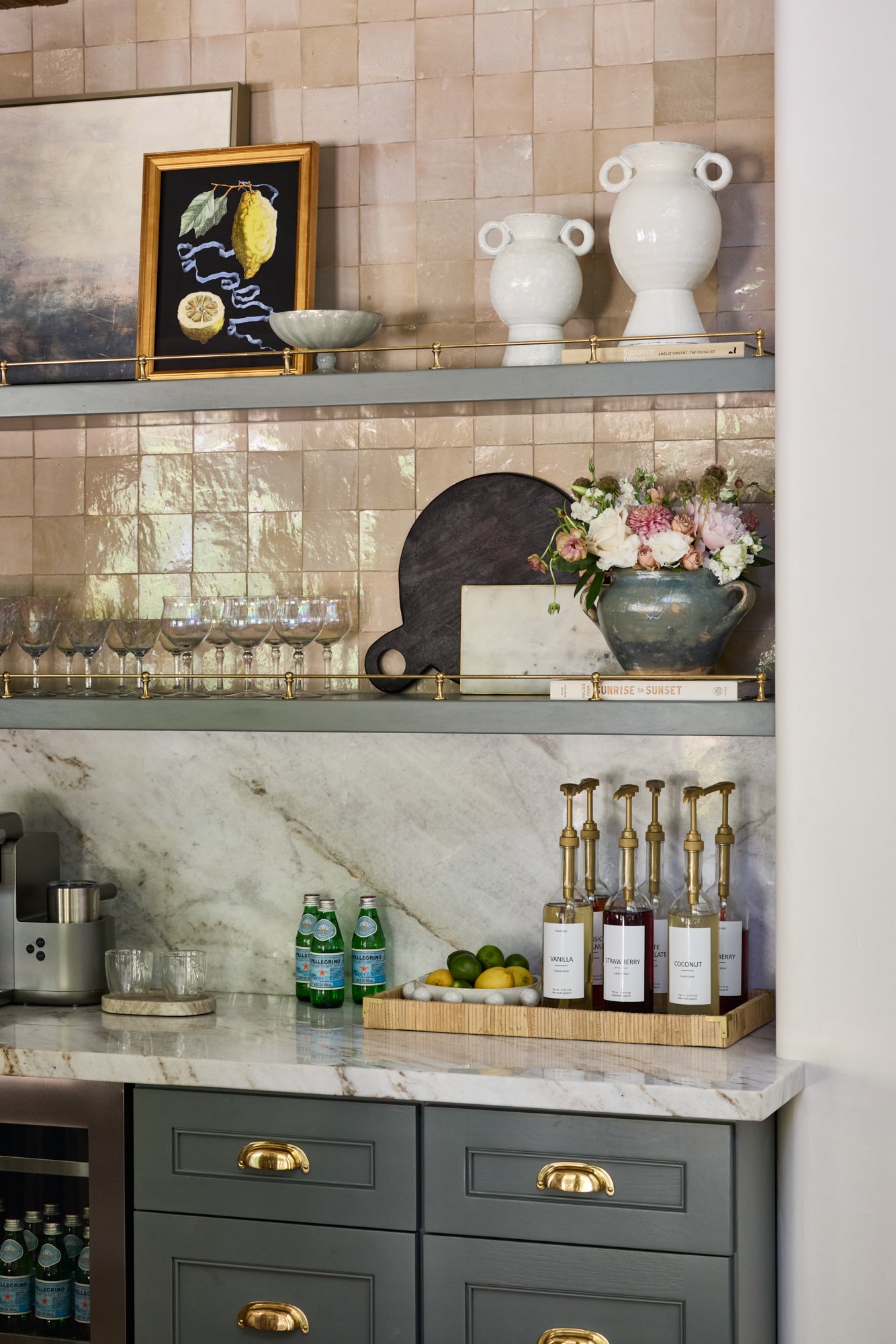 Decorative kitchen shelf with vases, framed artwork, flowers, glassware, and bottles of flavored syrup on a marble countertop.