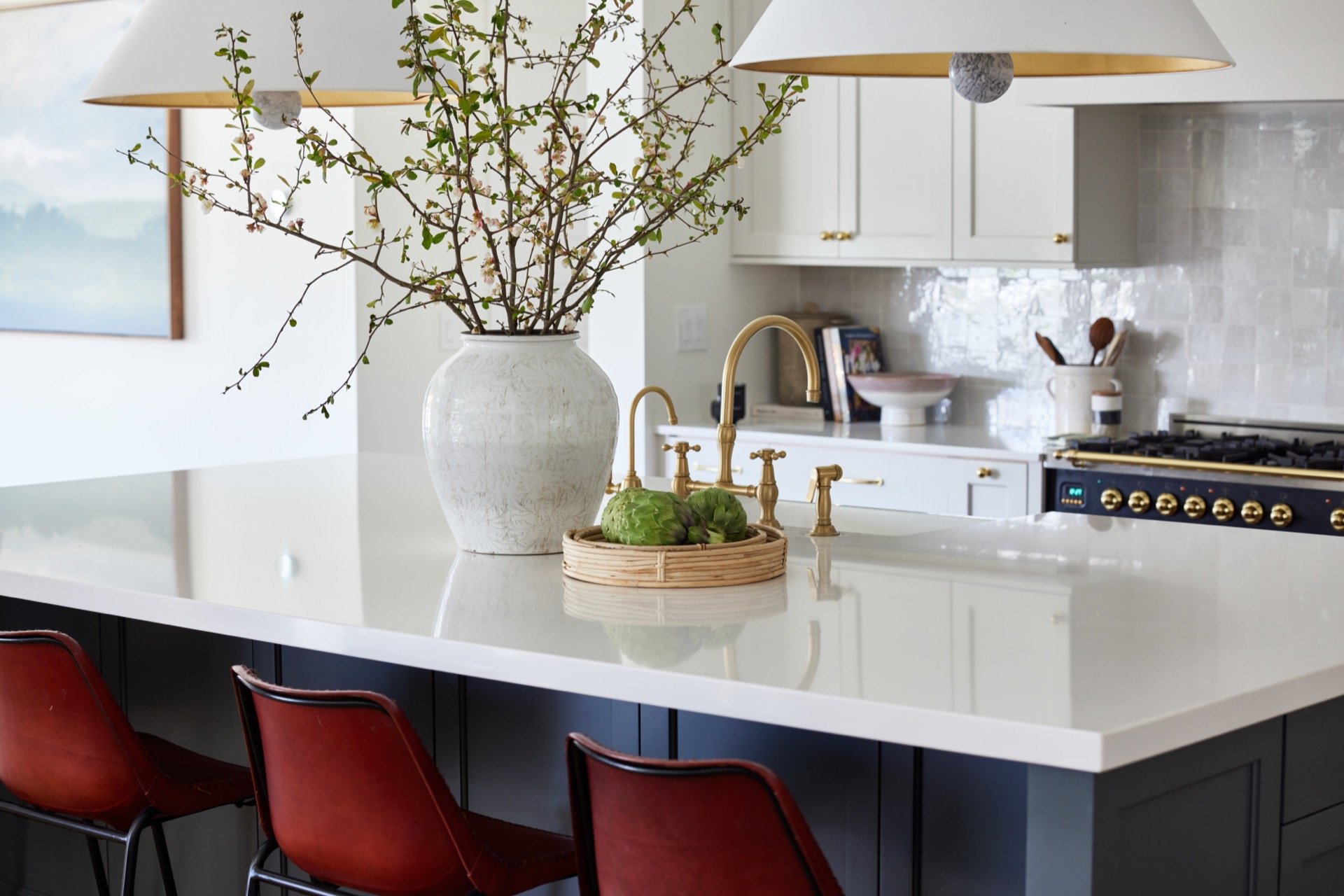Modern kitchen with a white countertop, brass faucet, potted plant, and wicker tray with artichokes. Red barstools and stove with brass accents are in the background.