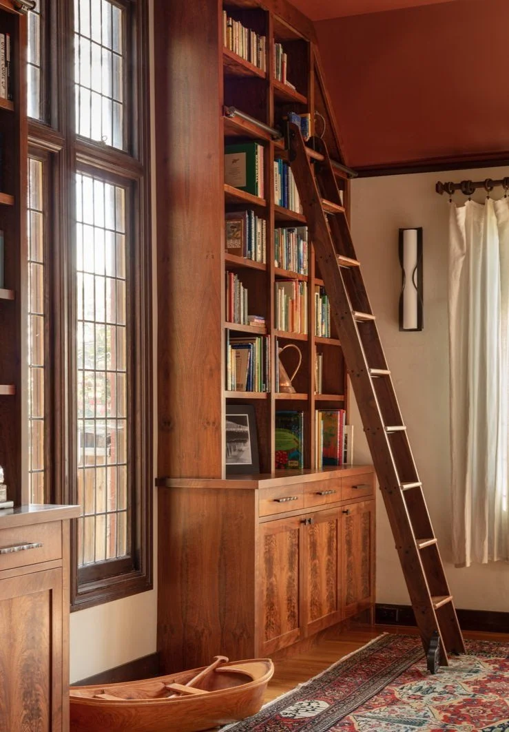 A wooden bookshelf with books and decorative items near a window with stained glass, a wooden ladder leaning against the bookshelf, and a wooden boat on the floor, in a cozy room with a patterned rug and curtains.
