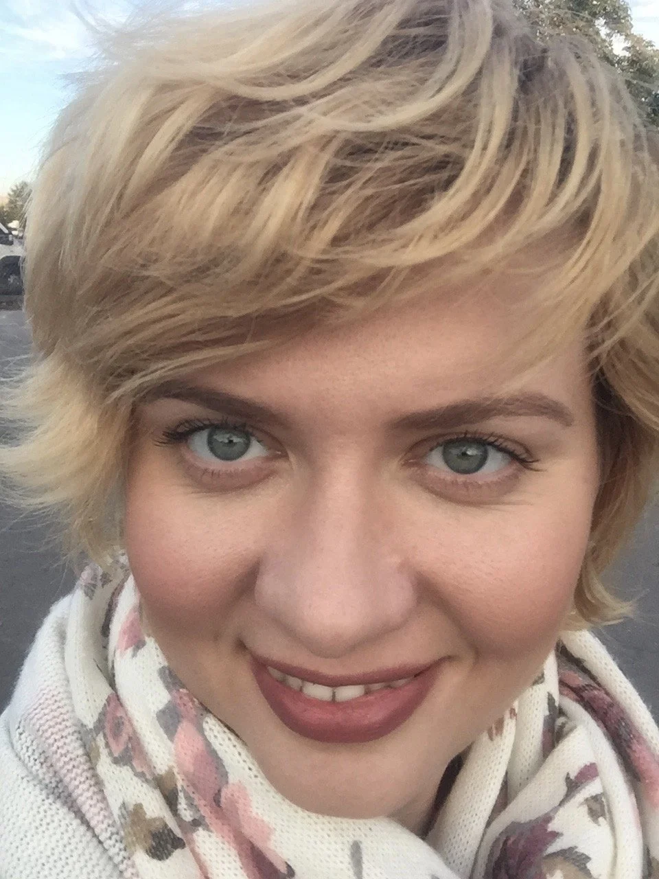 Close-up of a woman with short blonde hair, blue eyes, and wearing a floral scarf outdoors during daytime.