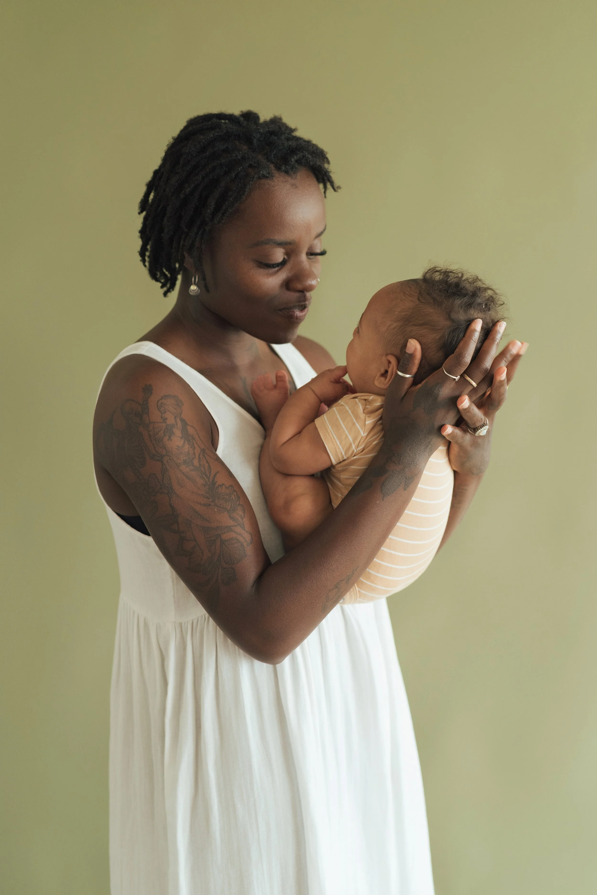 A woman with tattoos on her arm holding a baby close, both looking at each other against a plain green background.