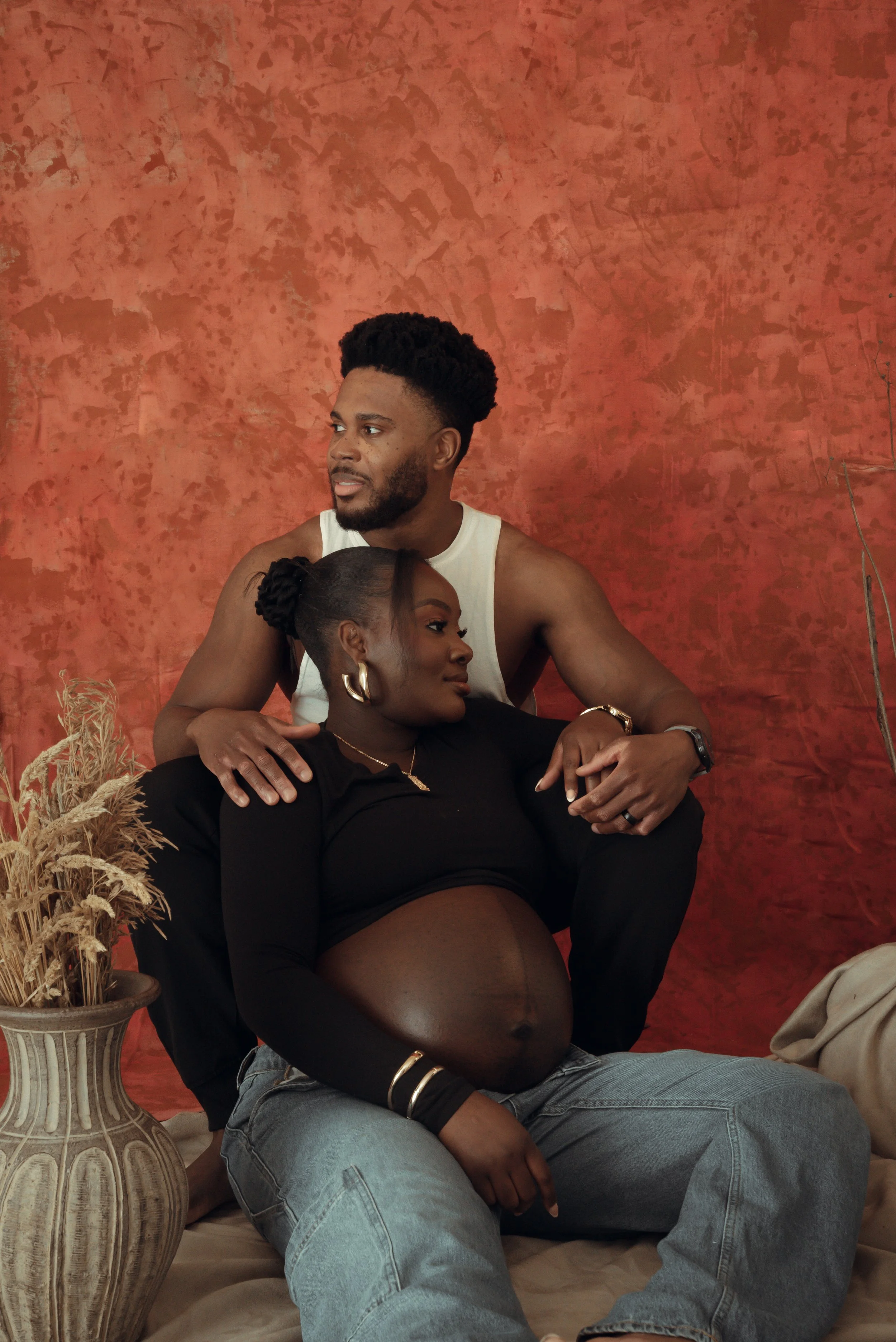 A pregnant woman sitting on the lap of a man, both relaxing with a textured reddish-brown backdrop behind them. The woman is dressed in black with gold hoop earrings, and the man is wearing a white tank top.