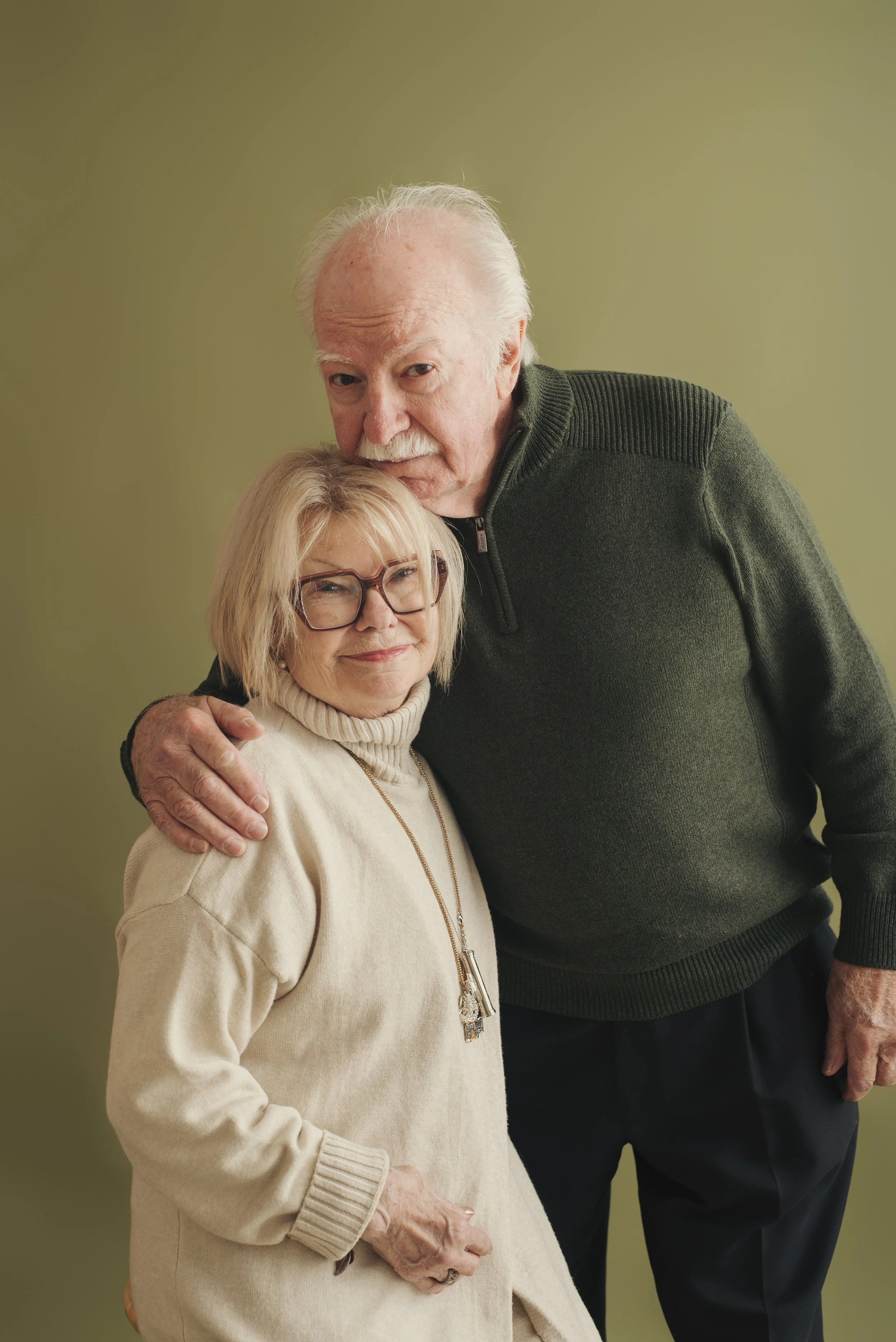 An elderly man with white hair and a mustache hugging an elderly woman with blonde hair and glasses against a green wall.