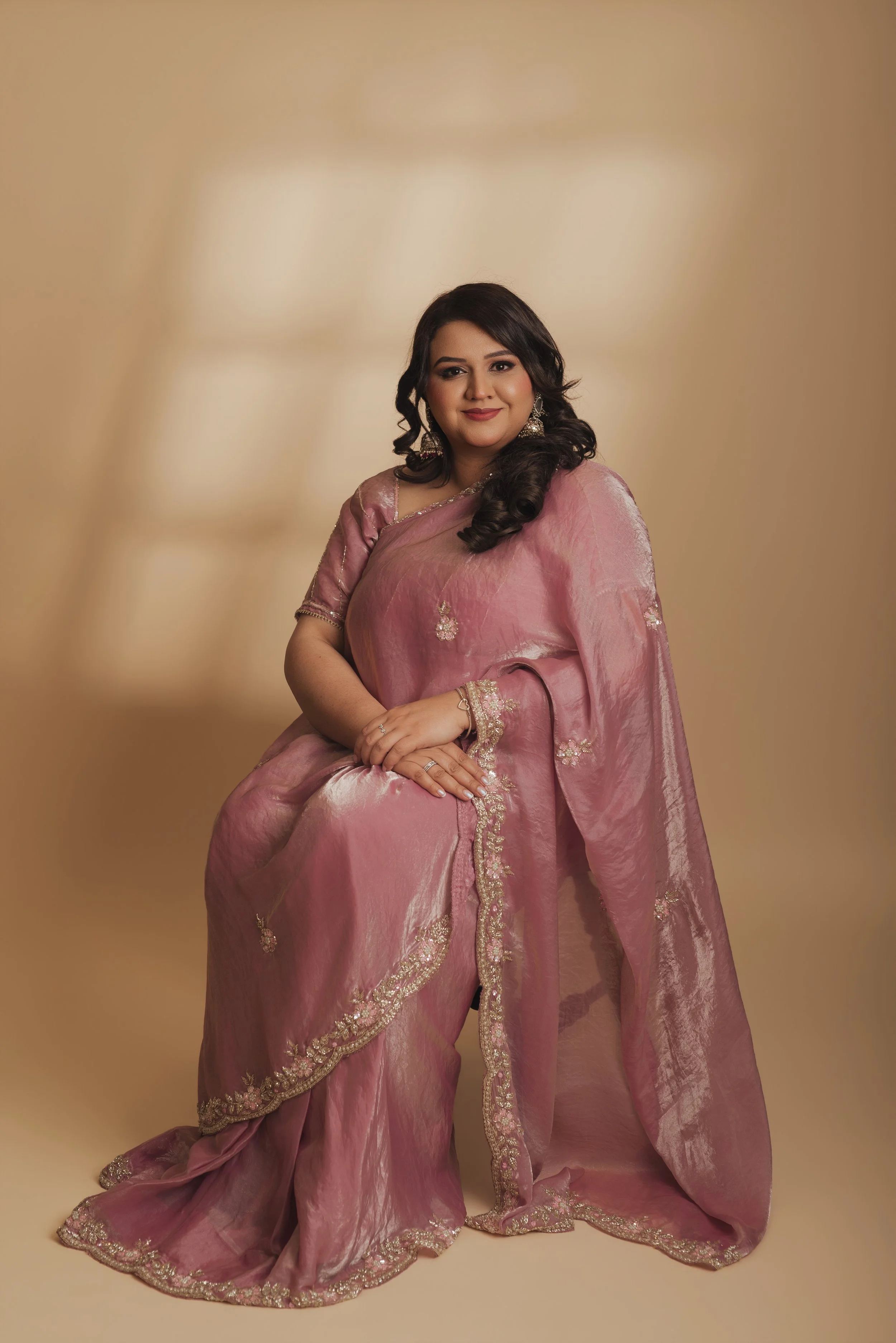 A woman with long dark hair wearing a pink traditional sari with gold embroidery, jewelry, and earrings, sitting against a beige background.