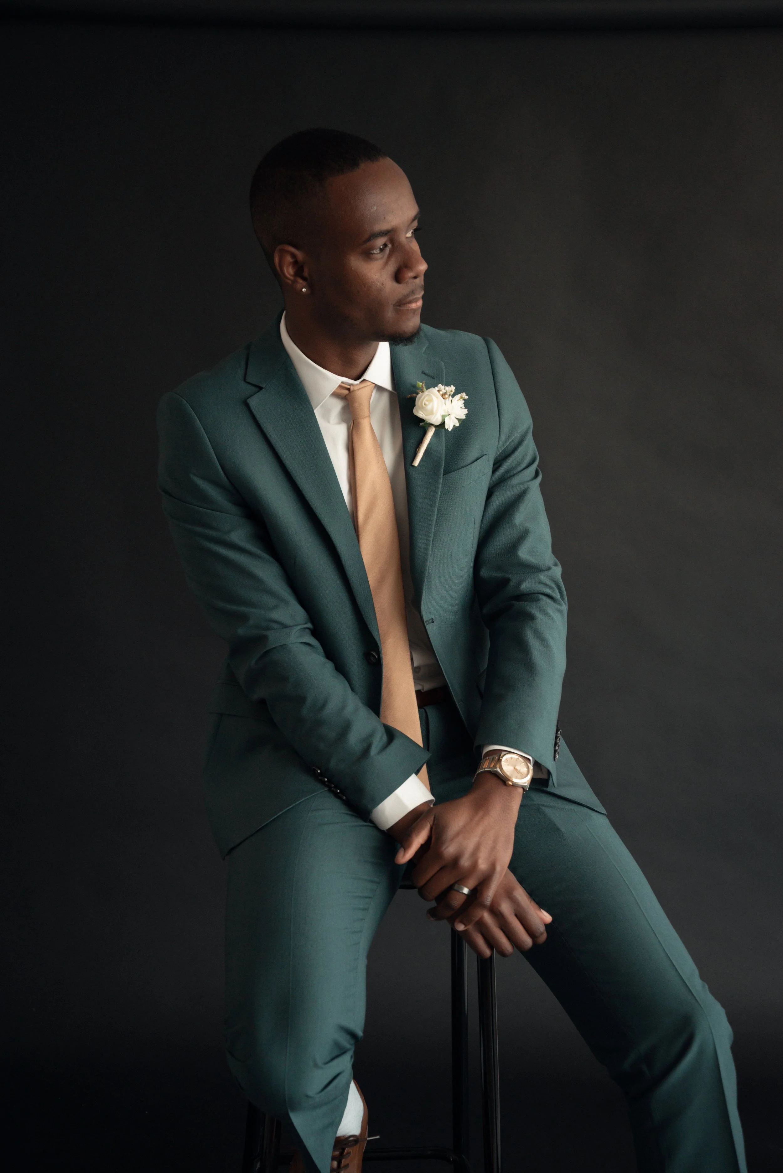 A man in a teal suit and beige tie sitting on a stool against a black background, with a white boutonniere on his lapel, looking thoughtfully to the side.