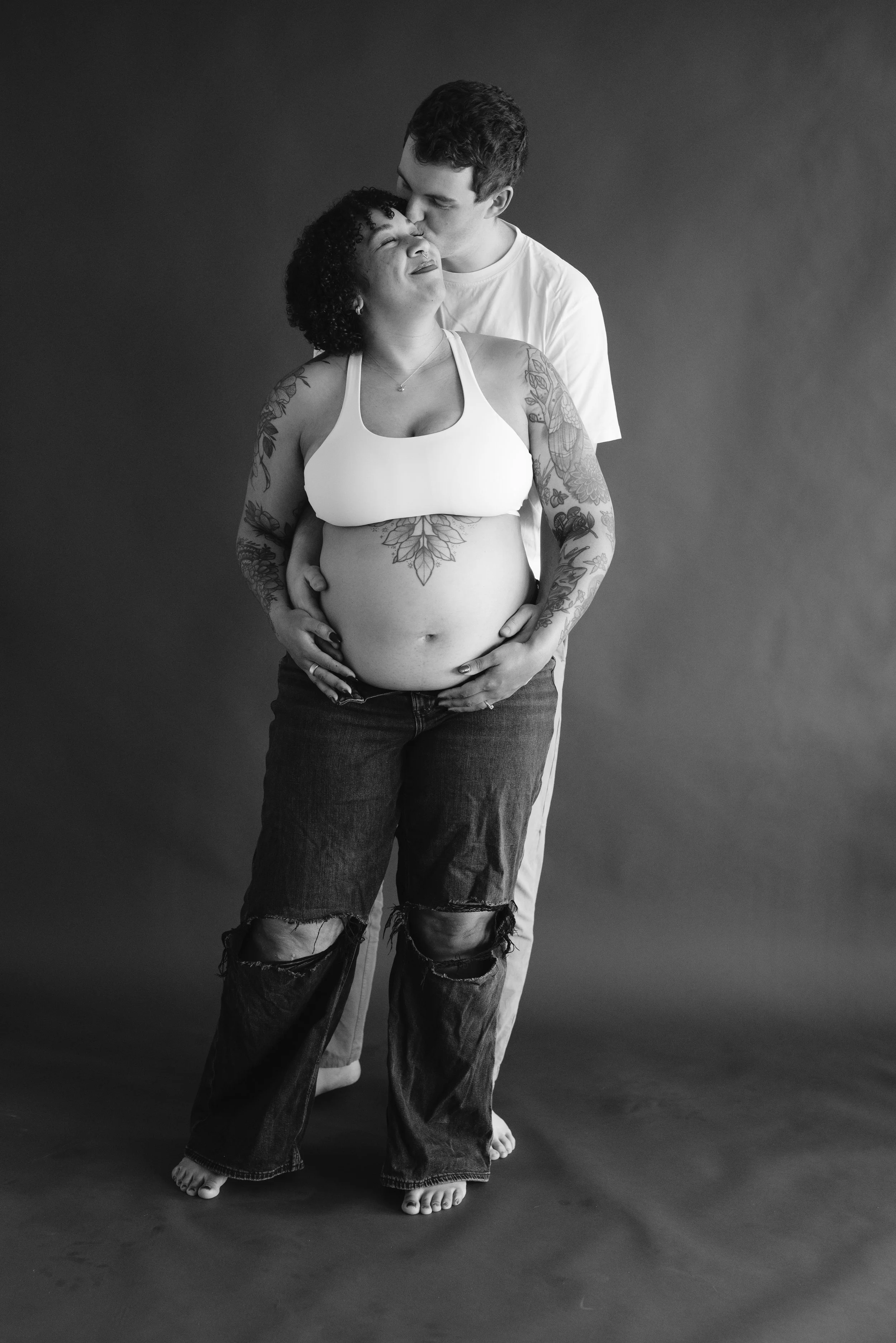 A black-and-white photo of a couple, with the man kissing the woman's forehead as she smiles with eyes closed. The woman has tattoos on her arms and torso, and they are holding each other. They are barefoot and standing on a plain surface against a s