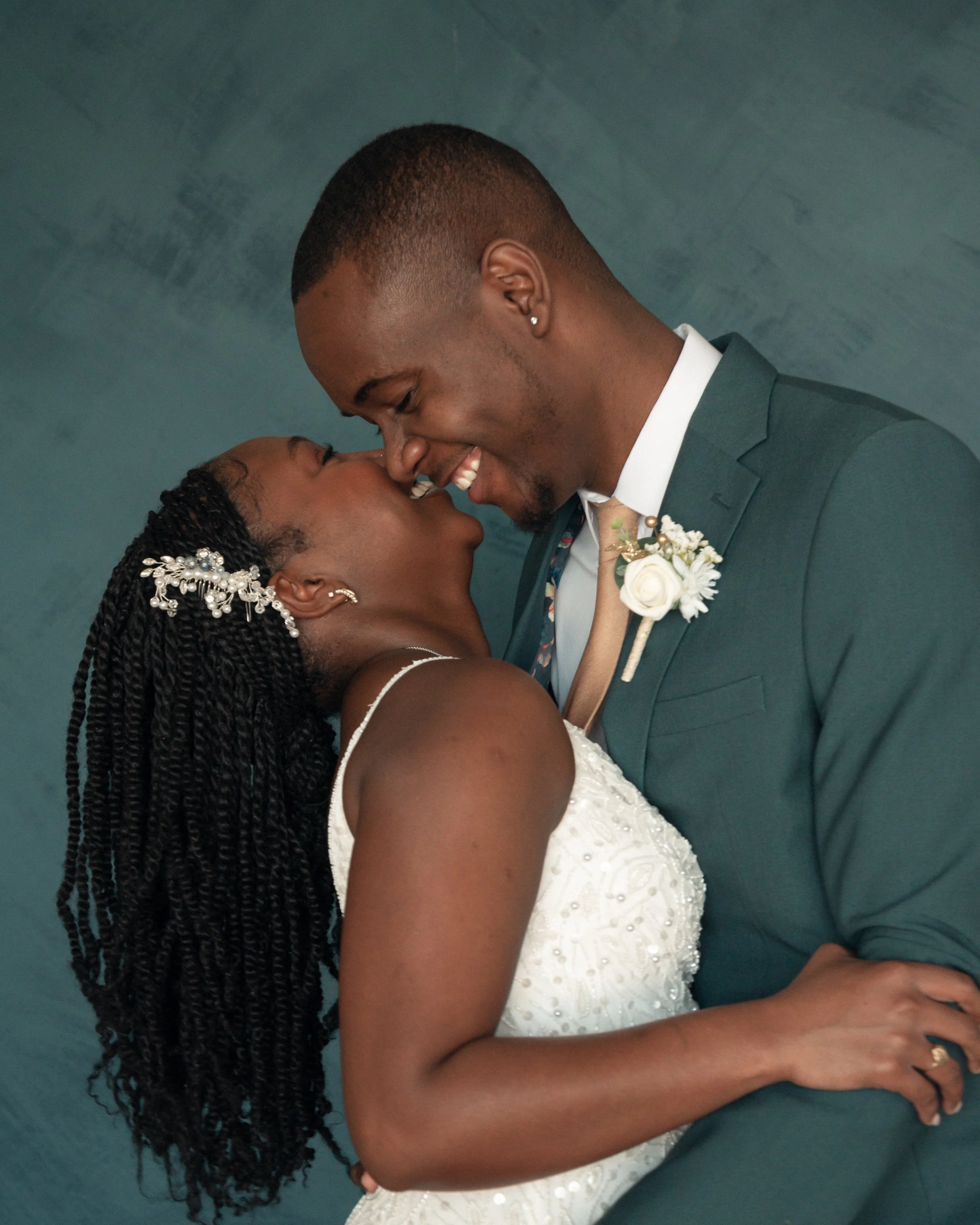 A close-up of a smiling bride and groom leaning their foreheads together during a wedding celebration. The bride has dark braided hair decorated with white flowers, and the groom is wearing a dark green suit with a boutonnière.